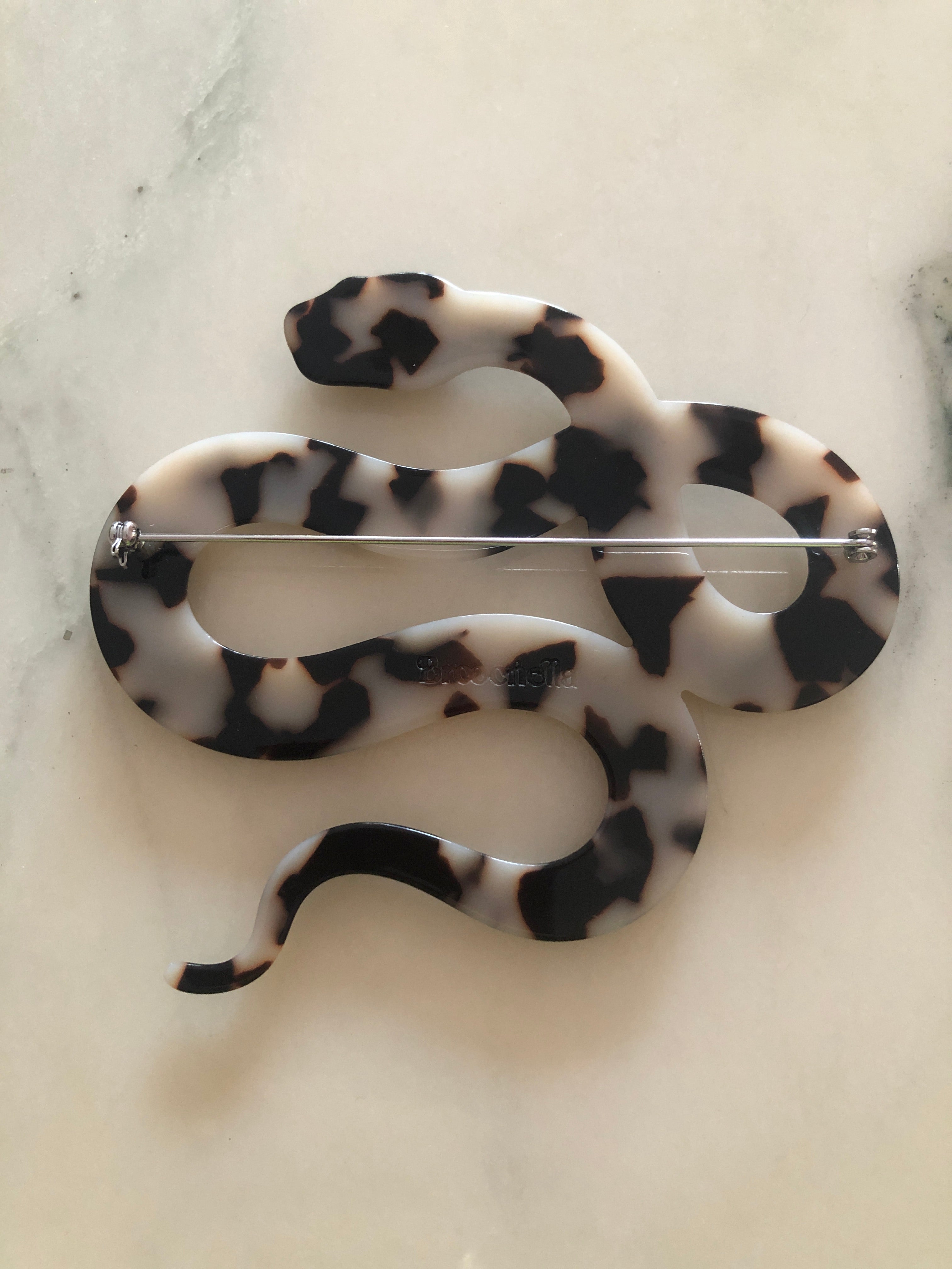 A Broochella Animalia Collection coiled snake brooch in black and white tortoiseshell Italian cellulose acetate with detailed scale texture measuring 9cm x 10cm, photographed on a white marble surface.