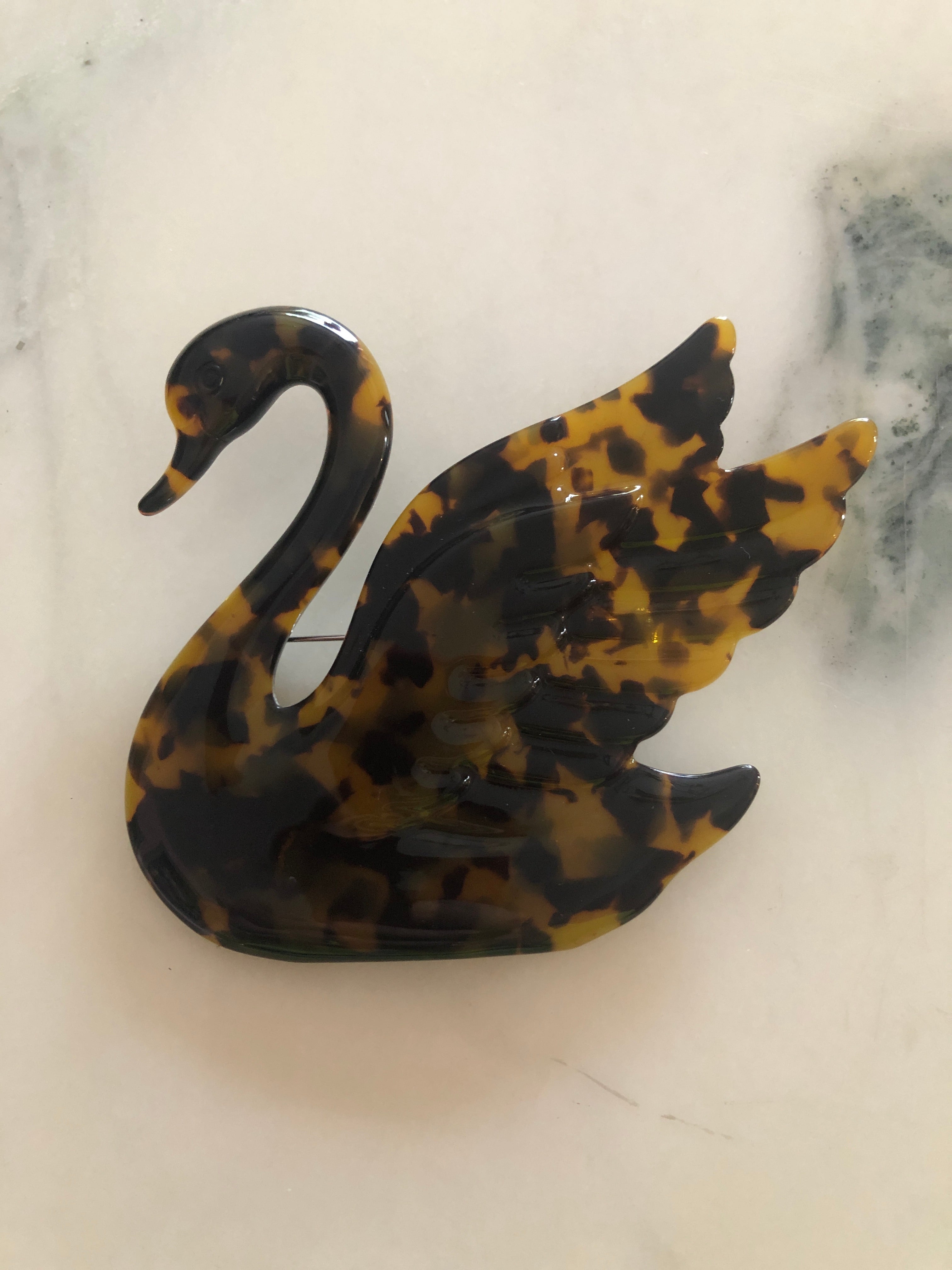 A Broochella Animalia Collection beige tortoiseshell swan brooch and men's lapel pin in Italian cellulose acetate. The background is white marble
