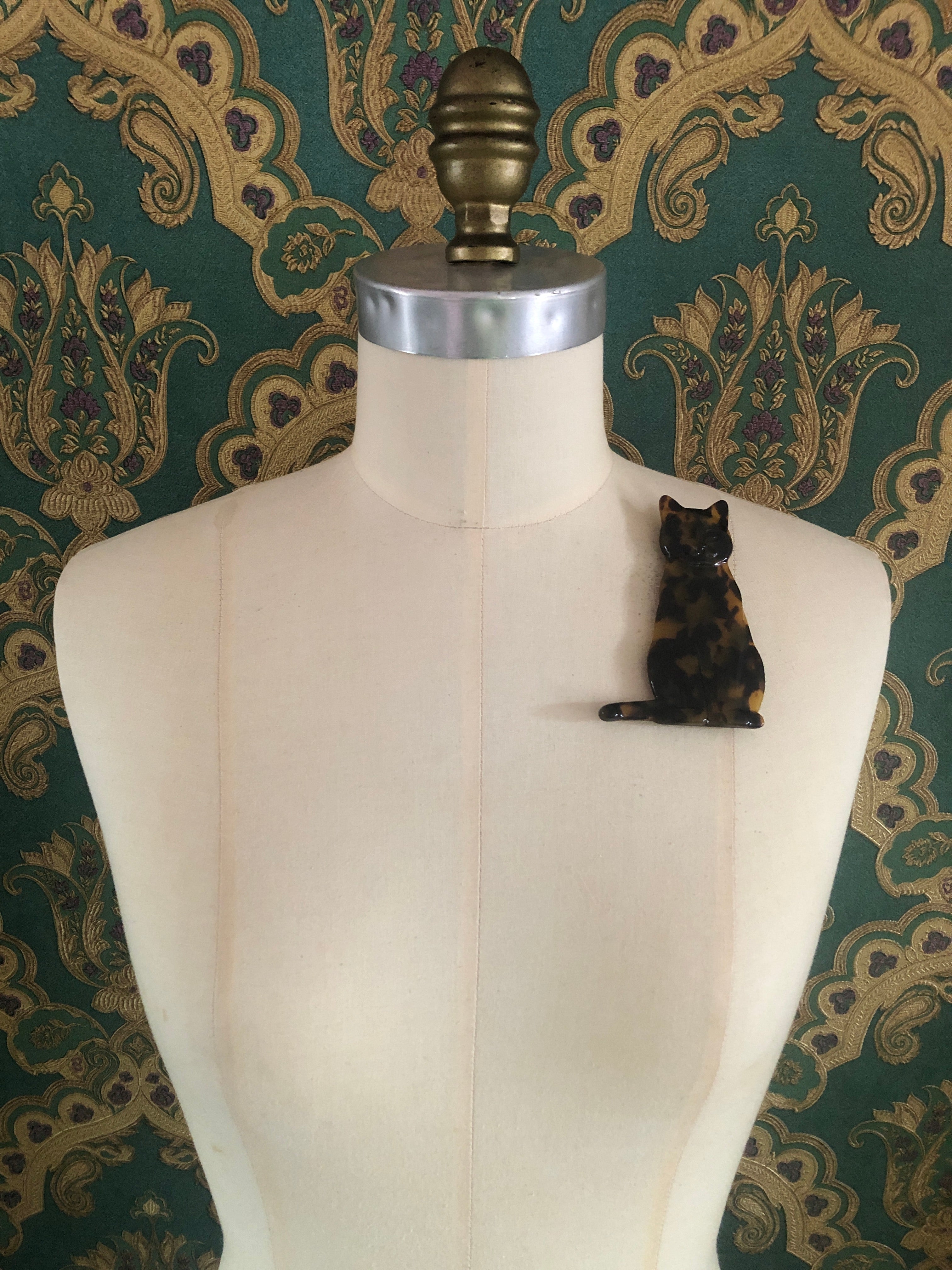 A Broochella Animalia Collection sitting cat brooch in brown tortoiseshell Italian cellulose acetate with sculpted face and curled tail detail measuring 10cm x 7cm, photographed on a white marble surface.