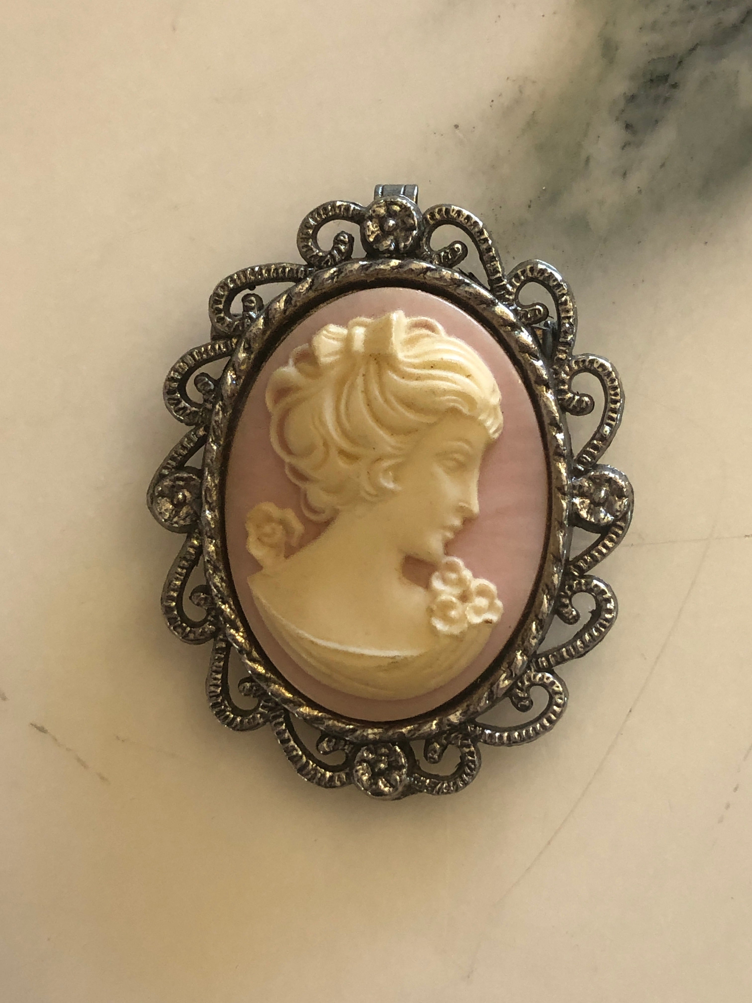 A 5.5cm x 4.5cm vintage cameo brooch featuring a carved cream classical portrait on a blush pink ground set within a silver-tone openwork filigree scrollwork frame, photographed on a white marble surface.