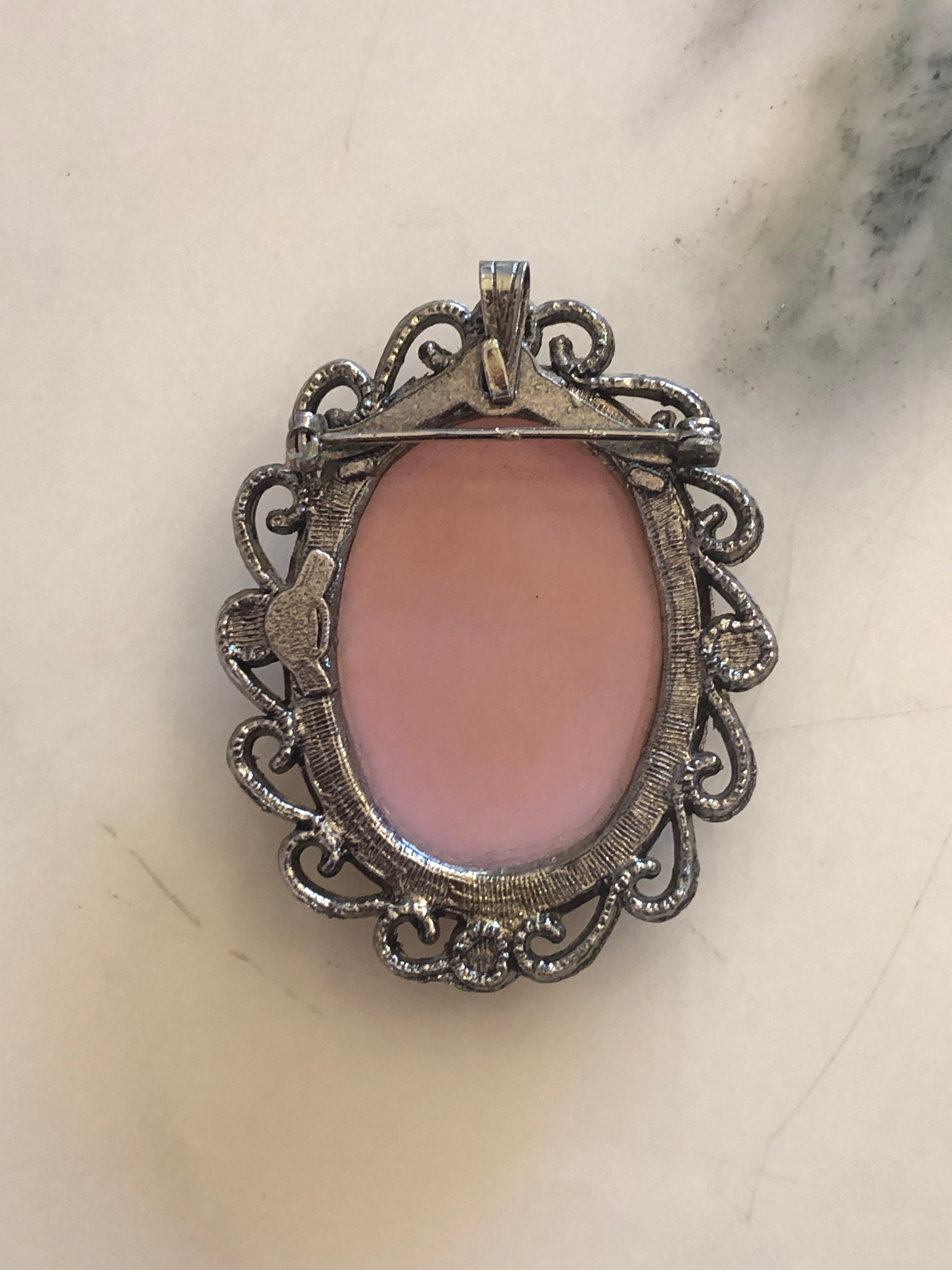 A 5.5cm x 4.5cm vintage cameo brooch featuring a carved cream classical portrait on a blush pink ground set within a silver-tone openwork filigree scrollwork frame, photographed on a white marble surface.