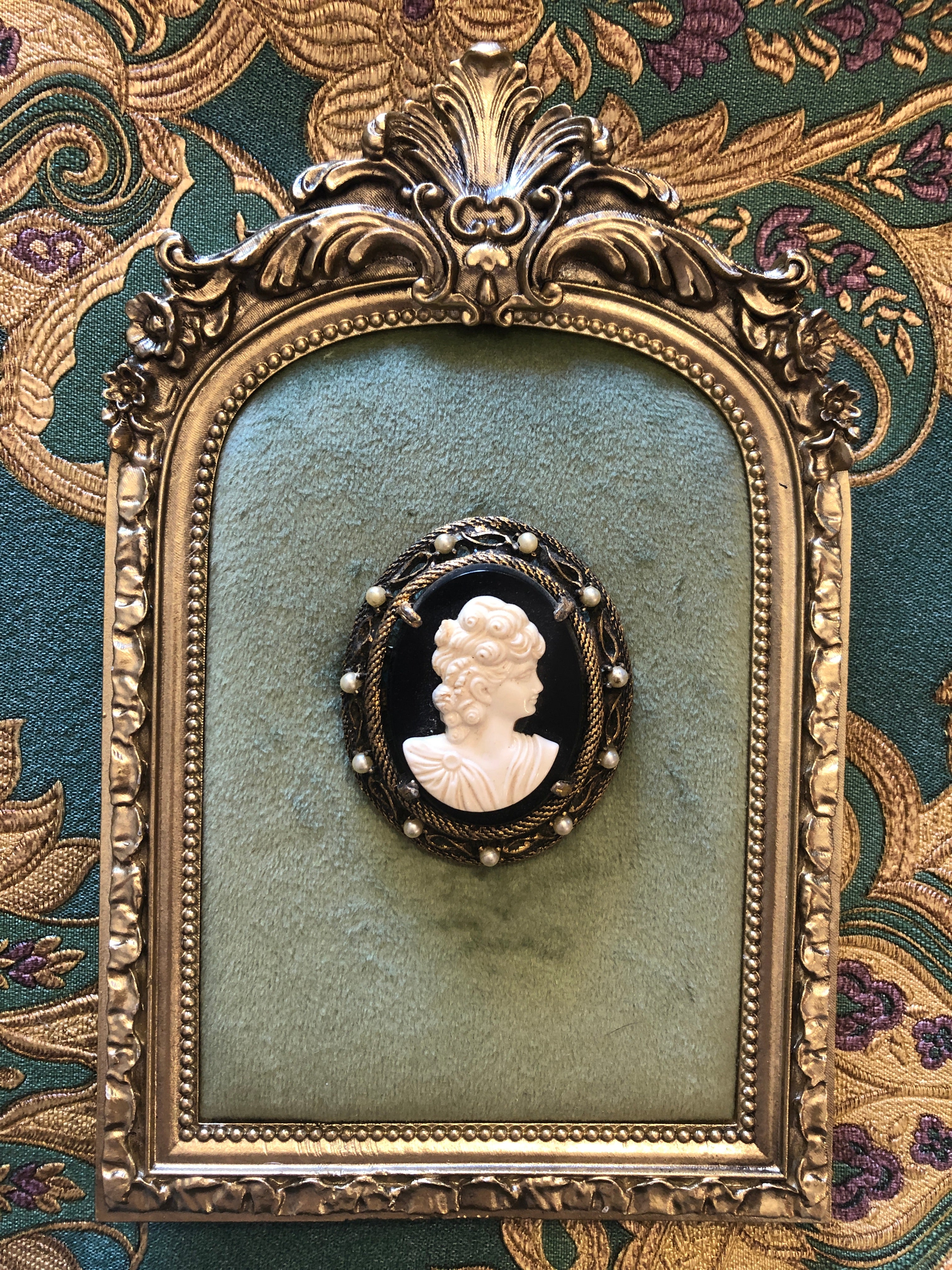 A 5.5cm x 4.5cm vintage cameo brooch featuring a carved white classical portrait on a deep black ground set within a gold-tone rope twist filigree frame with seed pearl accents, photographed on a white marble surface.