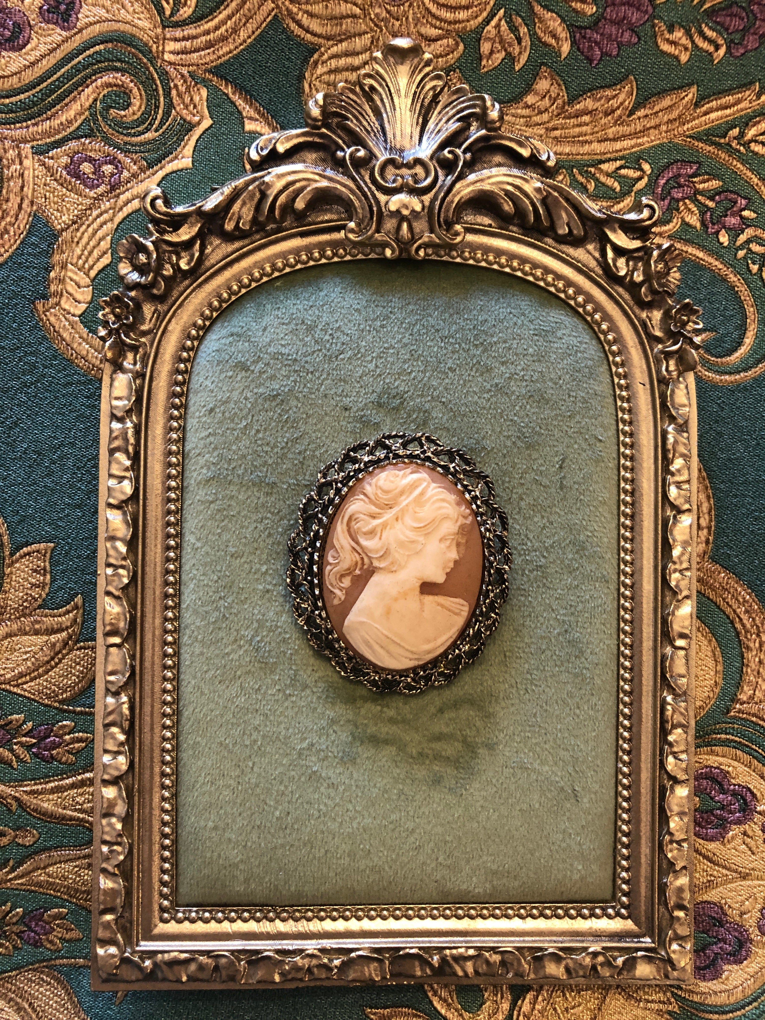 A 5cm x 4cm vintage cameo brooch featuring a carved classical portrait on a warm coral pink ground set within a wide gold-tone openwork rope twist filigree frame, photographed on a white marble surface.