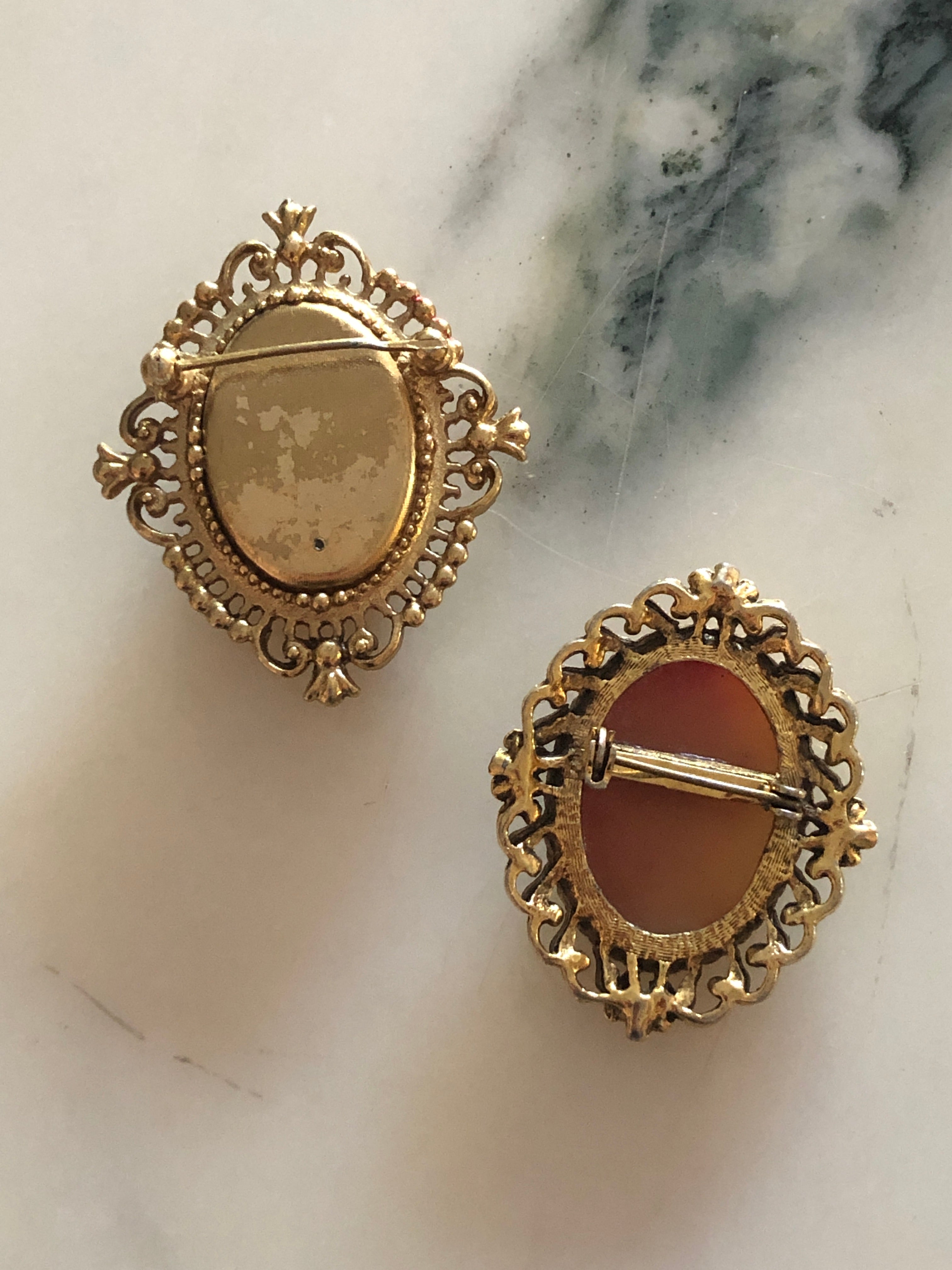 A set of two vintage cameo brooches featuring carved cream classical portraits on dusty rose pink grounds - a larger square antique gold-tone scrollwork frame measuring 5cm x 4cm and a smaller oval antique gold-tone floral frame measuring 4.5cm x 3.5cm, photographed on a white marble surface.
