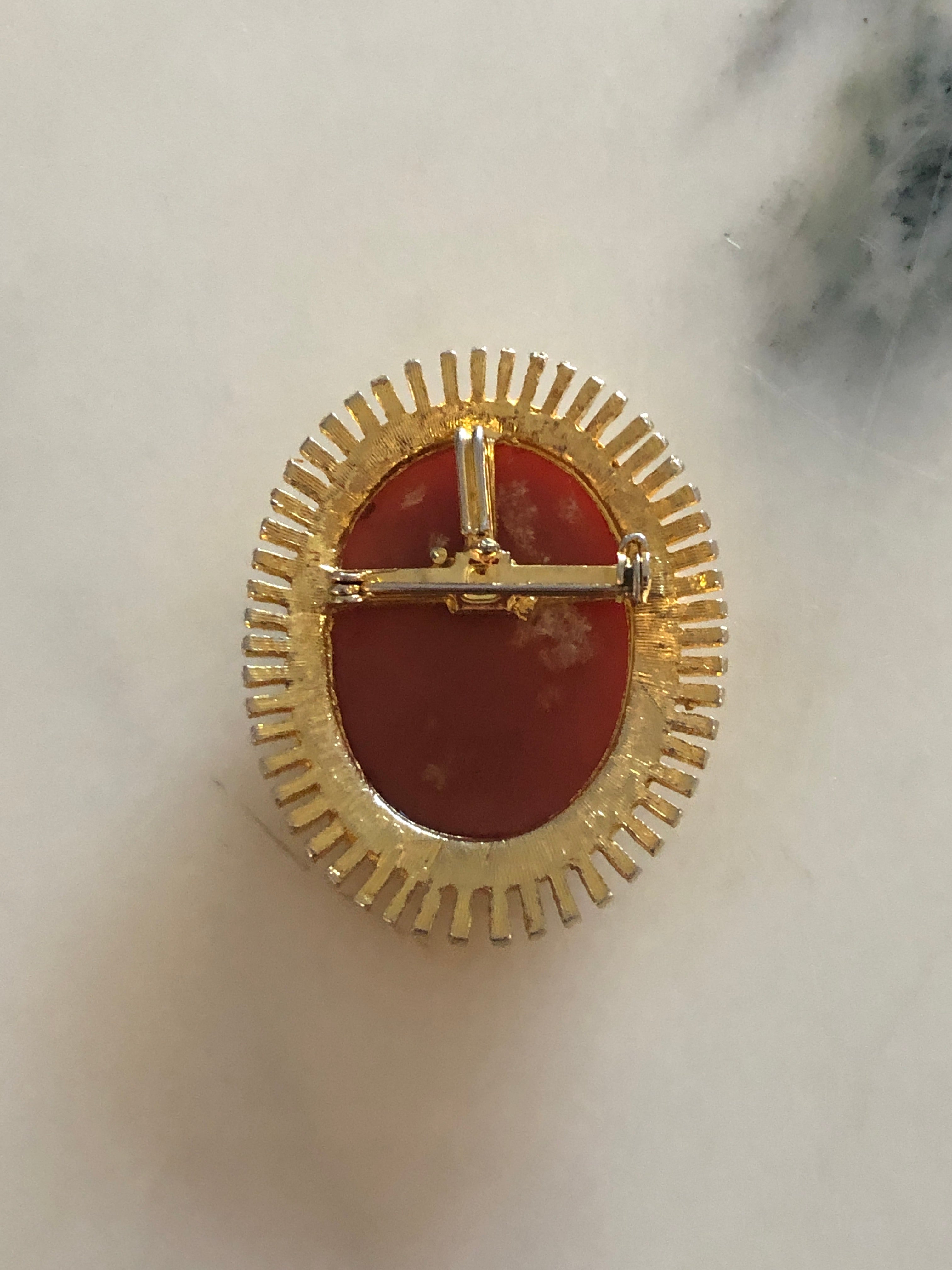 A 5cm x 4cm vintage cameo brooch featuring a carved cream classical portrait on a rich terracotta ground set within a gold-tone sunburst radiating bar frame, photographed on a white marble surface.