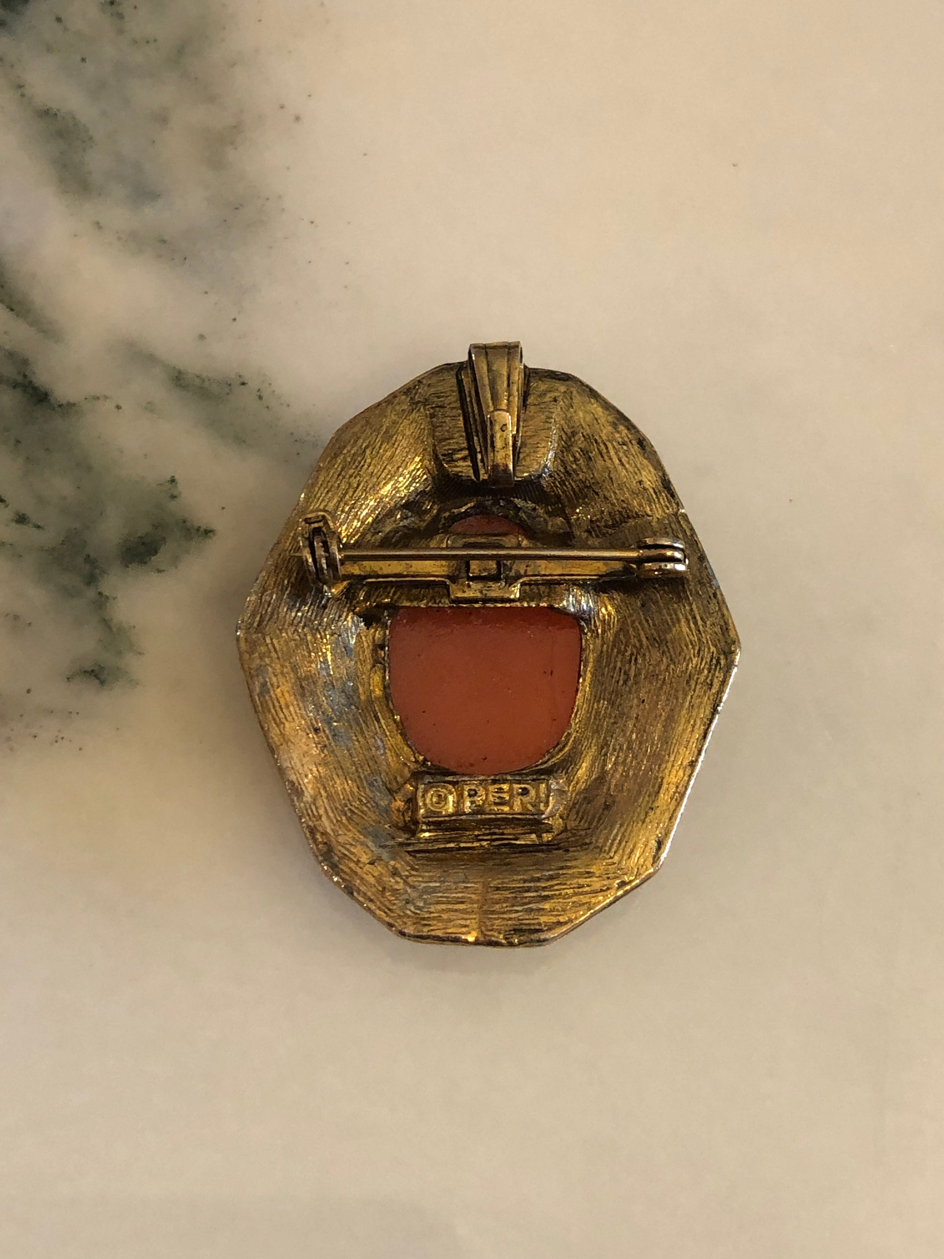 A 4cm x 3.5cm vintage cameo brooch featuring a carved white classical portrait on a warm terracotta ground set within an octagonal antique gold-tone rope twist and repousse scrollwork frame, photographed on a white marble surface.
