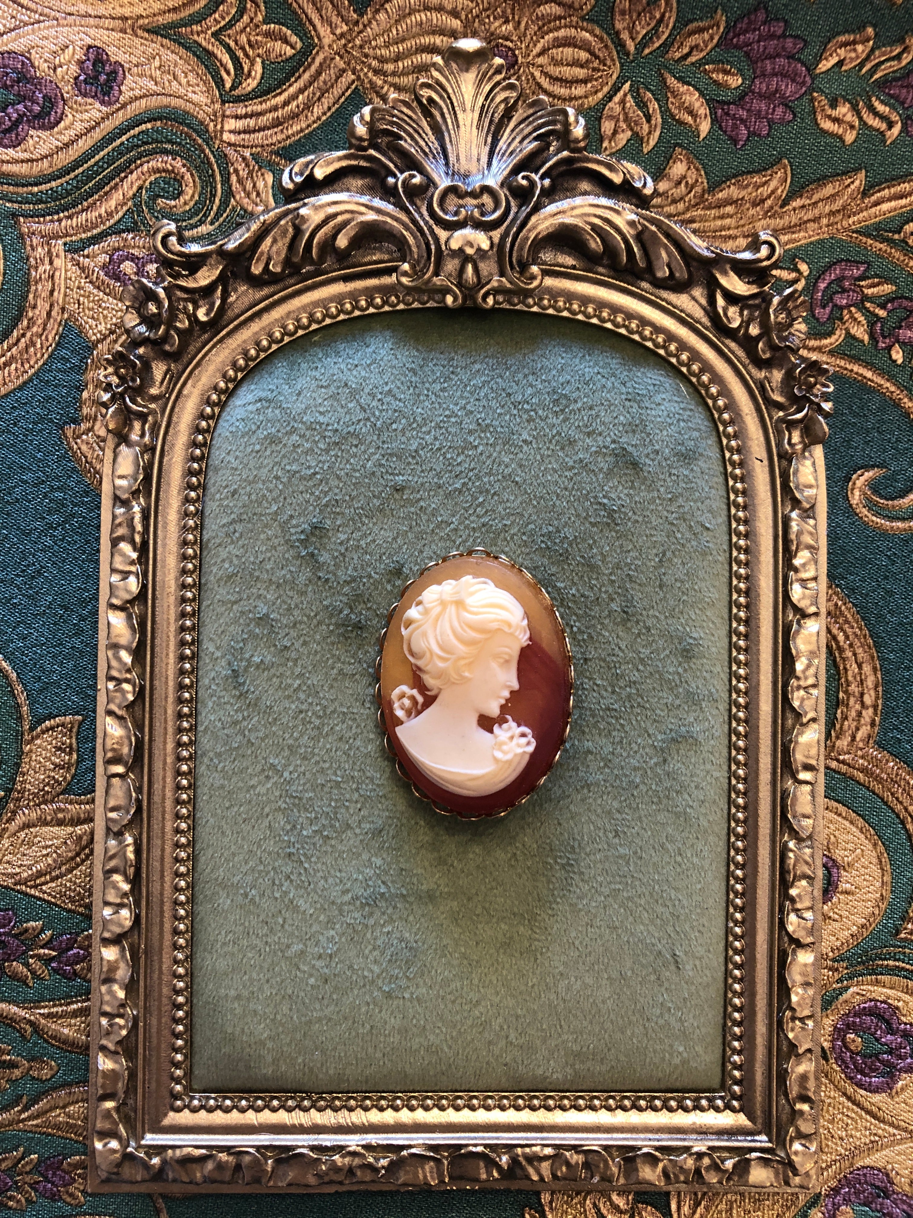 A 4cm x 3cm vintage cameo brooch featuring a carved cream classical portrait with bow and rose detail on a rich amber brown ground set within a gold-tone scalloped edge oval frame, photographed on a white marble surface.
