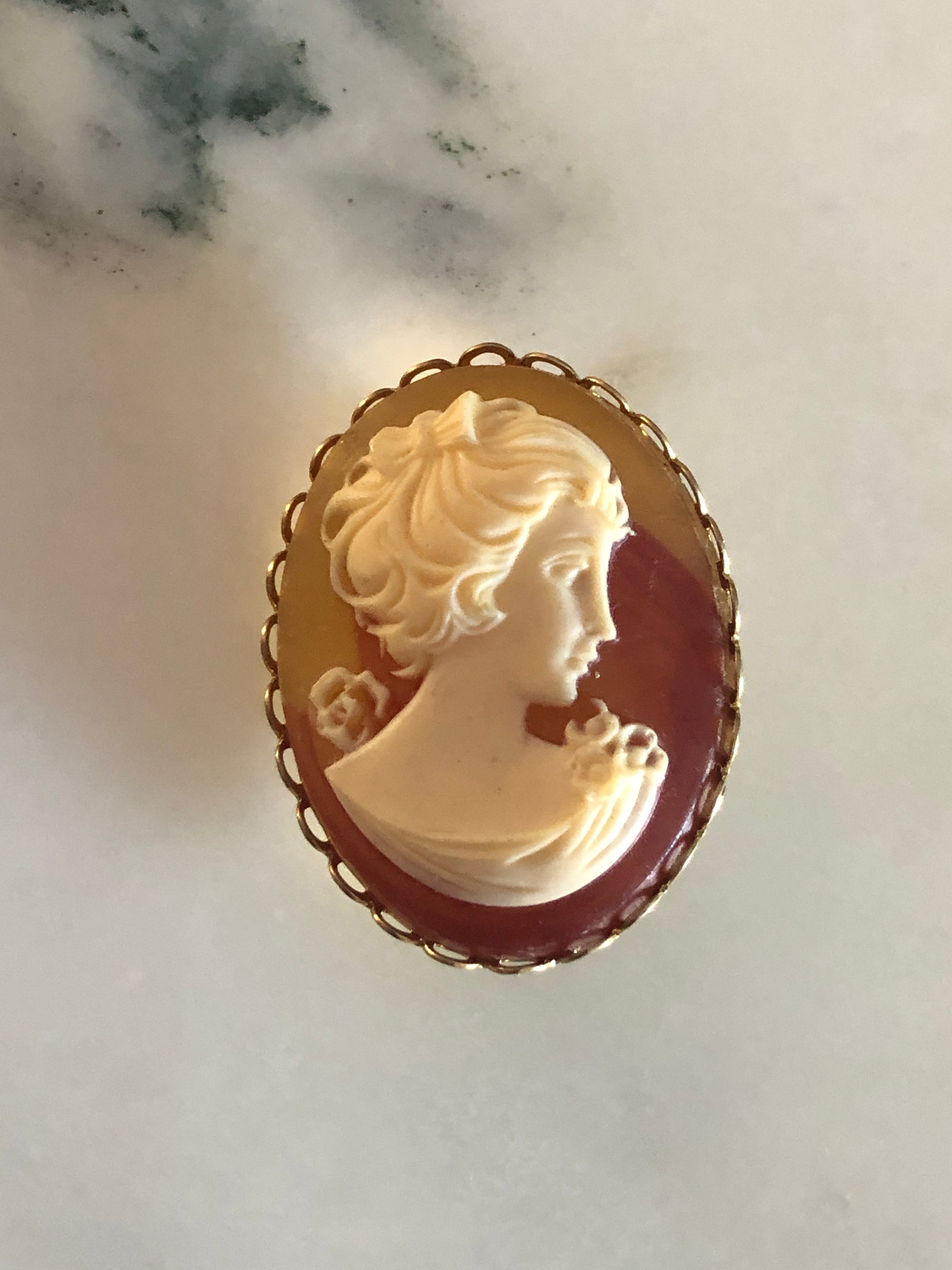 A 4cm x 3cm vintage cameo brooch featuring a carved cream classical portrait with bow and rose detail on a rich amber brown ground set within a gold-tone scalloped edge oval frame, photographed on a white marble surface.