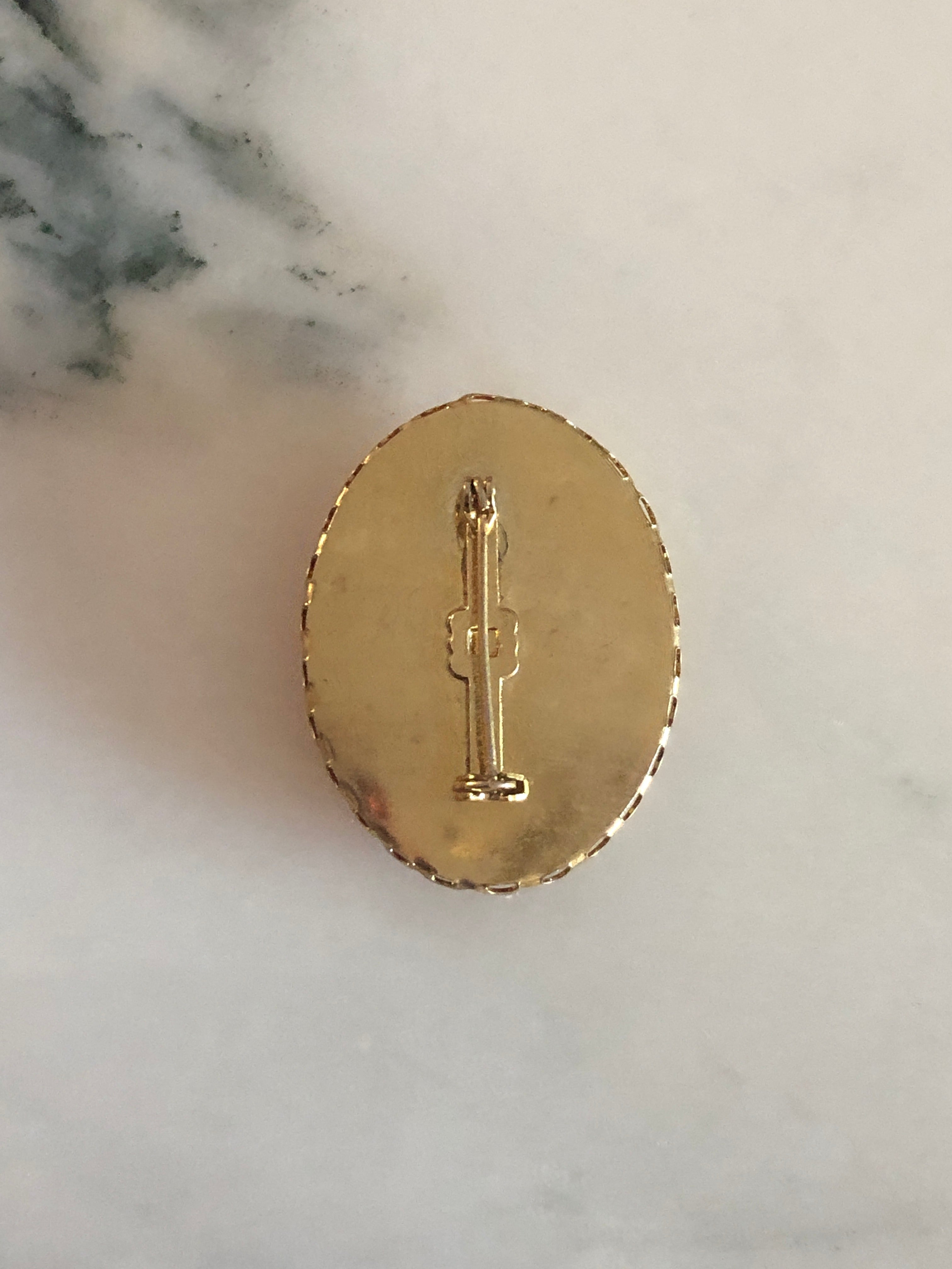 A 4cm x 3cm vintage cameo brooch featuring a carved cream classical portrait with bow and rose detail on a rich amber brown ground set within a gold-tone scalloped edge oval frame, photographed on a white marble surface.