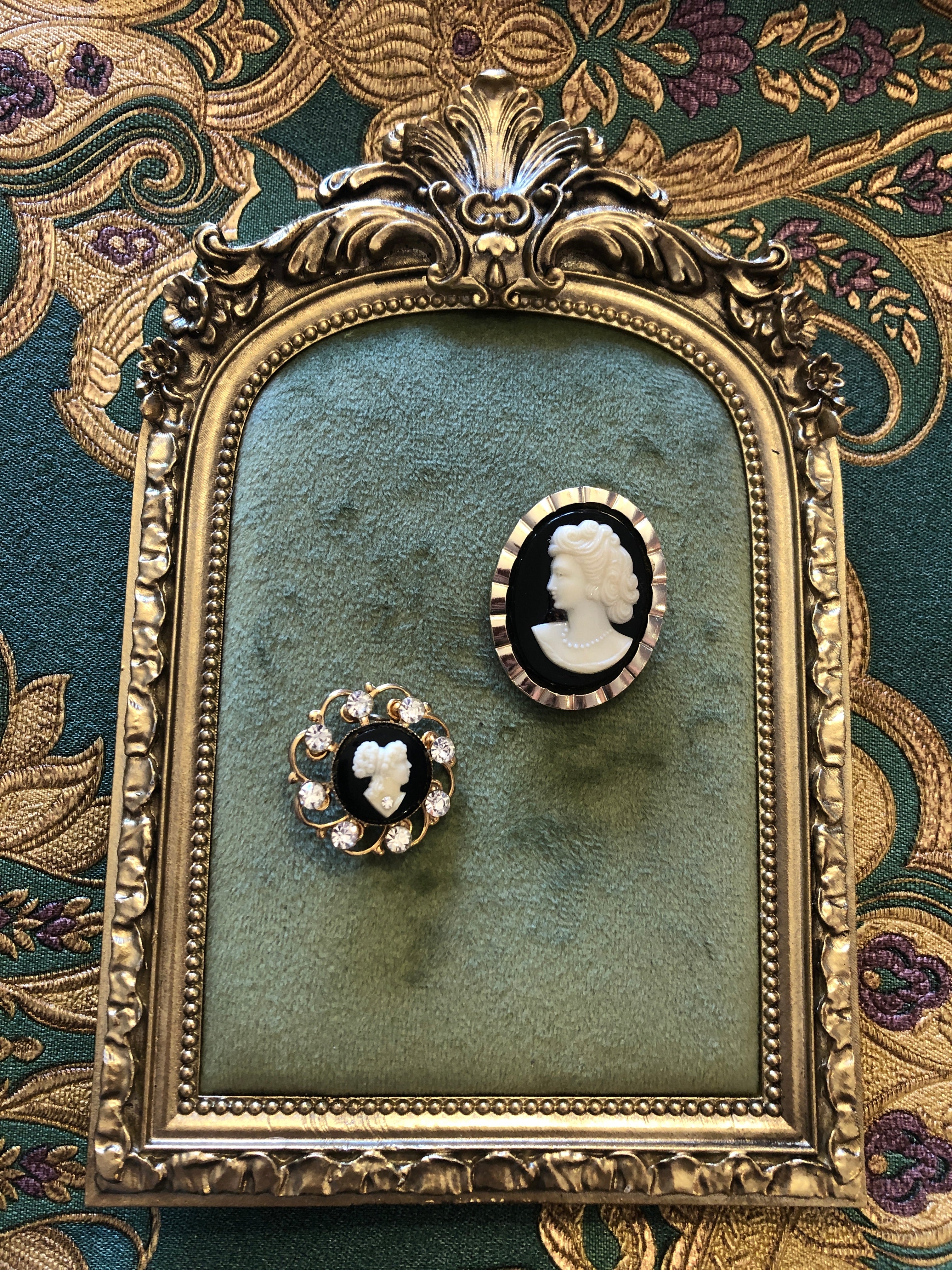 A vintage black and white cameo set comprising a gold-tone ridged oval tie clip measuring 3.5cm x 2.5cm and a circular gold-tone openwork scrollwork brooch measuring 2.5cm, both featuring carved white classical portraits on a deep black ground, photographed on a white marble surface.