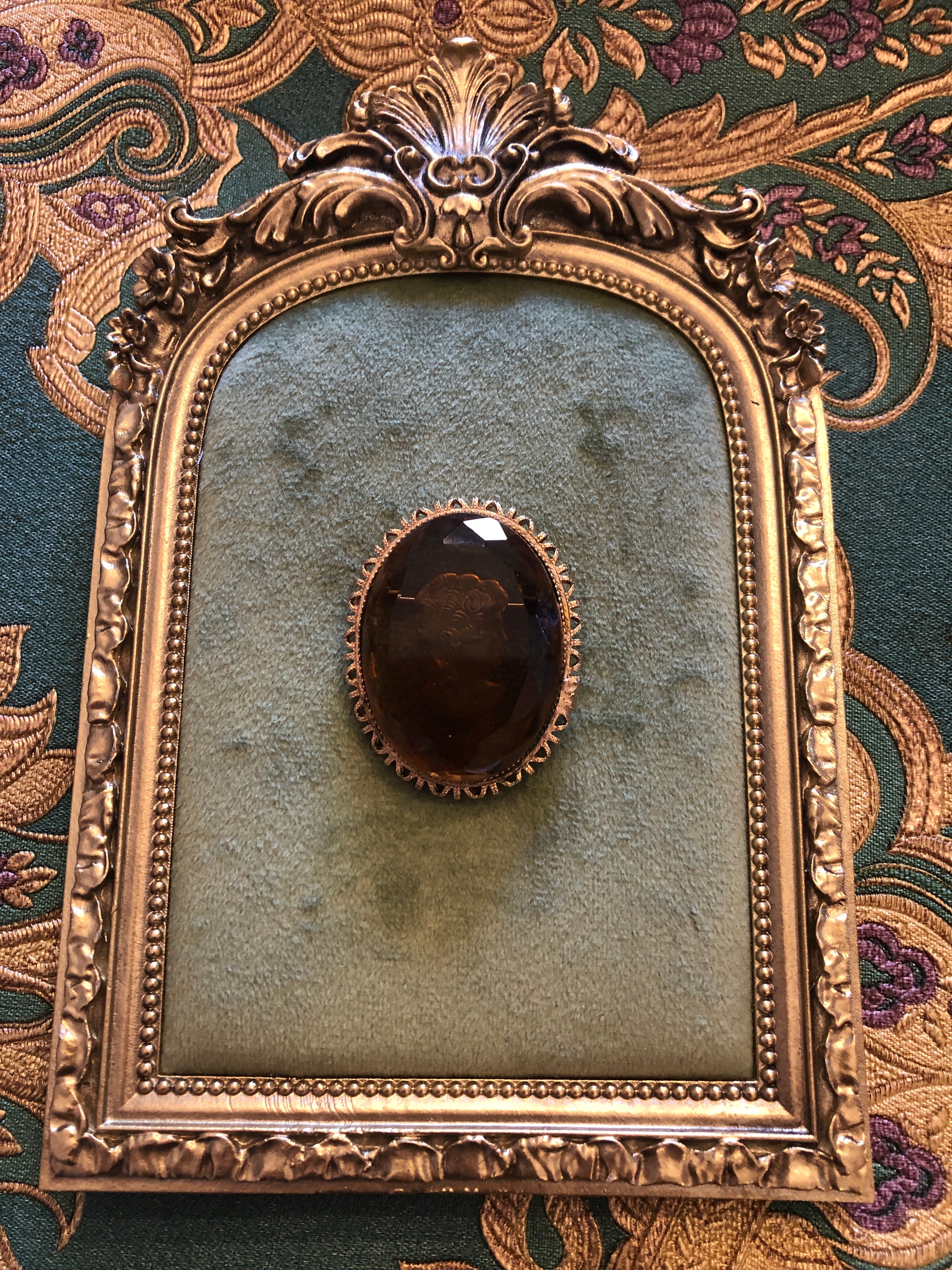 A 4.5cm x 3.5cm vintage intaglio cameo brooch featuring a classical portrait carved within a large faceted smoky glass stone set within a gold-tone castellated filigree edge oval frame, photographed on a white marble surface.