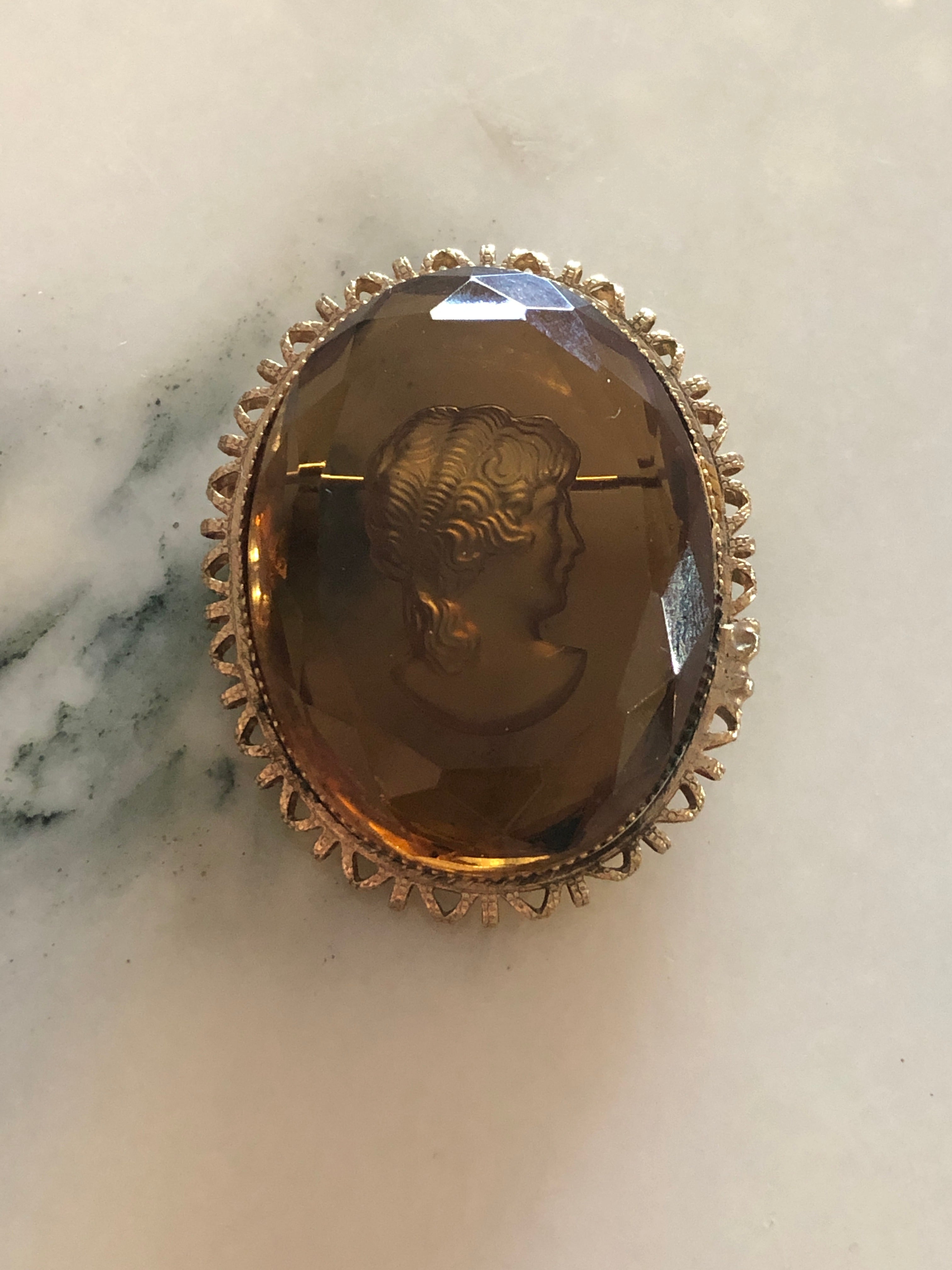 A 4.5cm x 3.5cm vintage intaglio cameo brooch featuring a classical portrait carved within a large faceted smoky glass stone set within a gold-tone castellated filigree edge oval frame, photographed on a white marble surface.