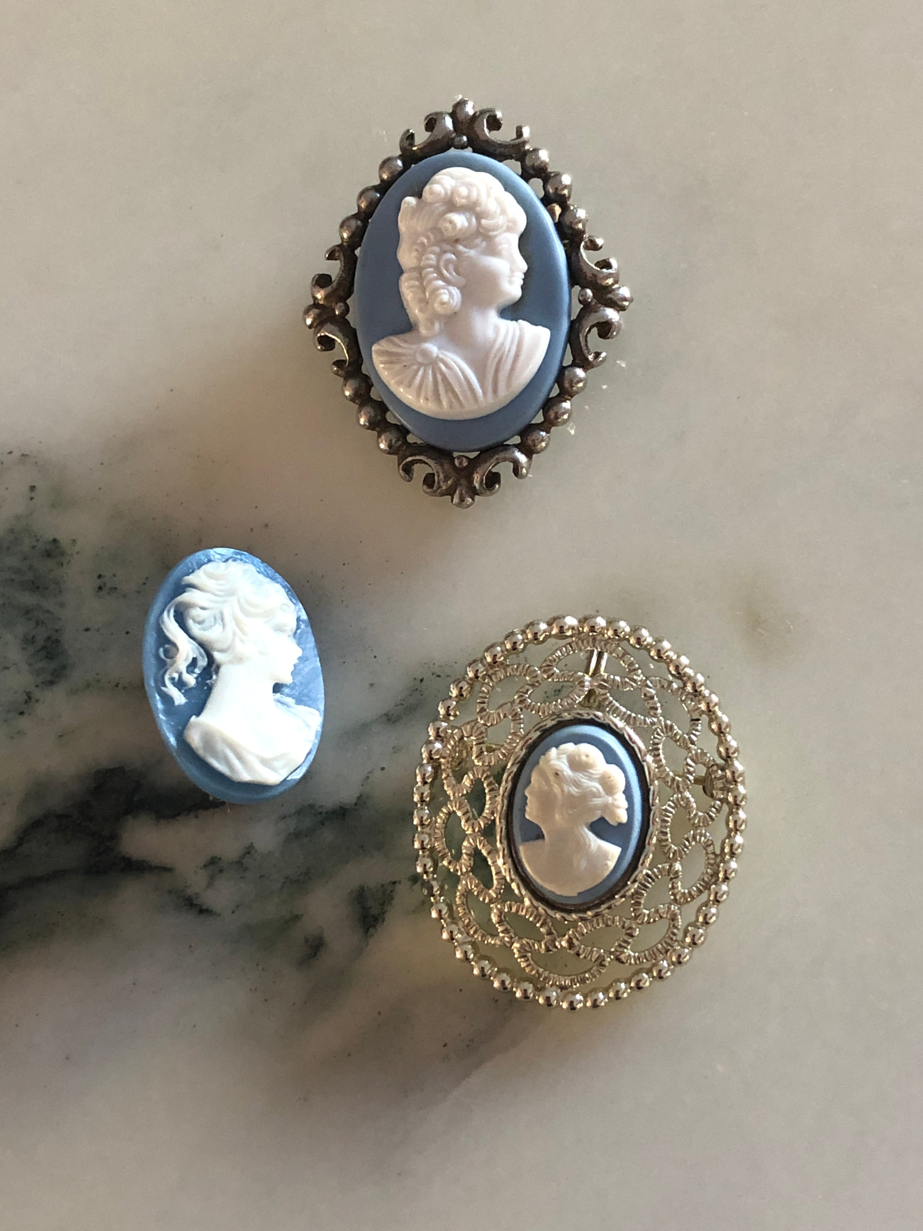 A set of three vintage blue and white cameo brooches featuring carved white classical portraits on powder blue grounds - in a gold-tone circular filigree frame, a silver-tone scrollwork frame and an unframed oval, ranging from 2.5cm to 4cm, photographed on a white marble surface.