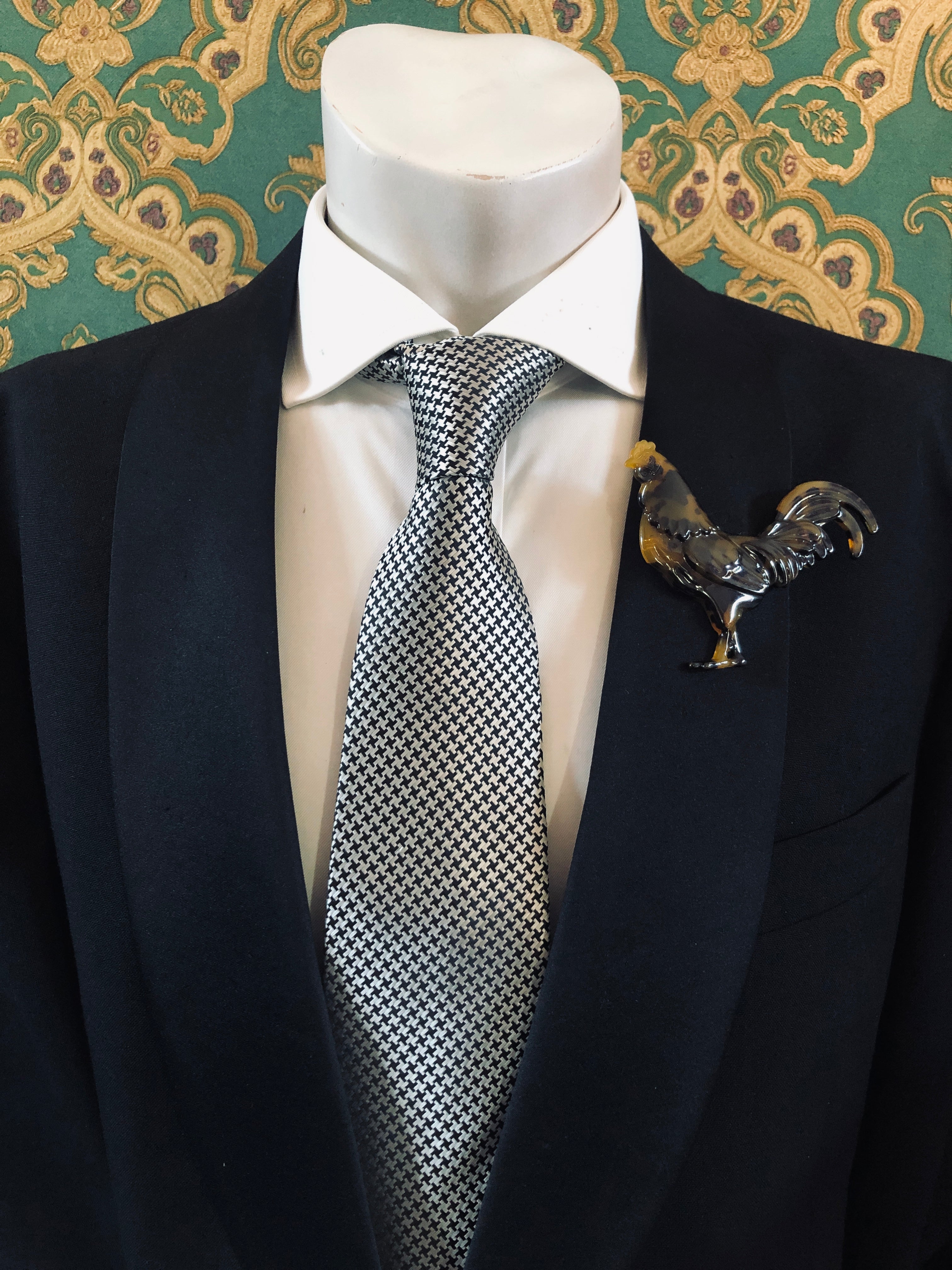 A Broochella Animalia Collection beige tortoiseshell rooster brooch and men's lapel pin in Italian cellulose acetate worn on the lapel of a black suit jacket with a white shirt and houndstooth tie, styled on a mannequin against a green embroidered fabric background.