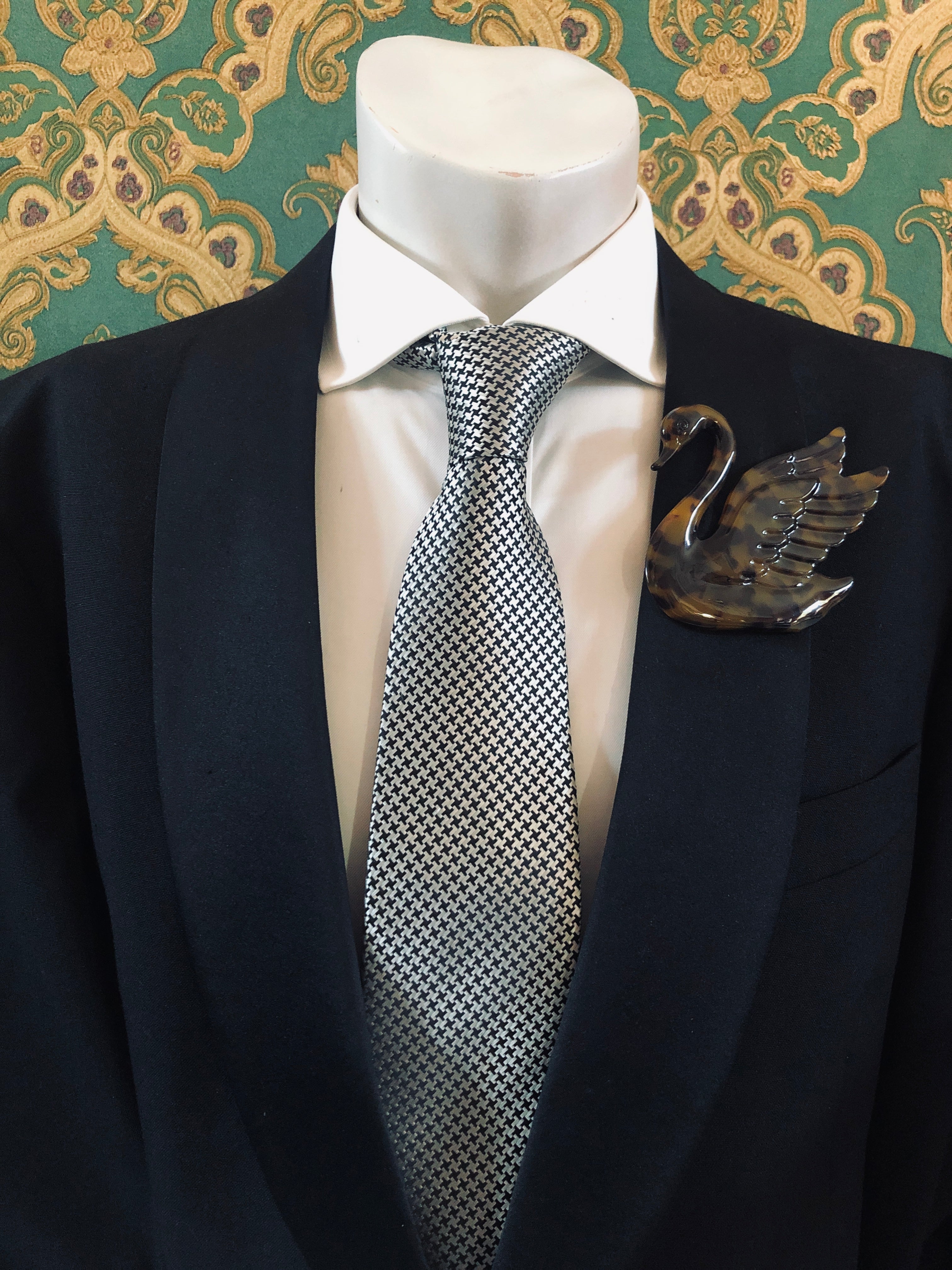 A Broochella Animalia Collection beige tortoiseshell swan brooch and men's lapel pin in Italian cellulose acetate worn on the lapel of a black suit jacket with a white shirt and houndstooth tie, styled on a mannequin against green and gold damask wallpaper.