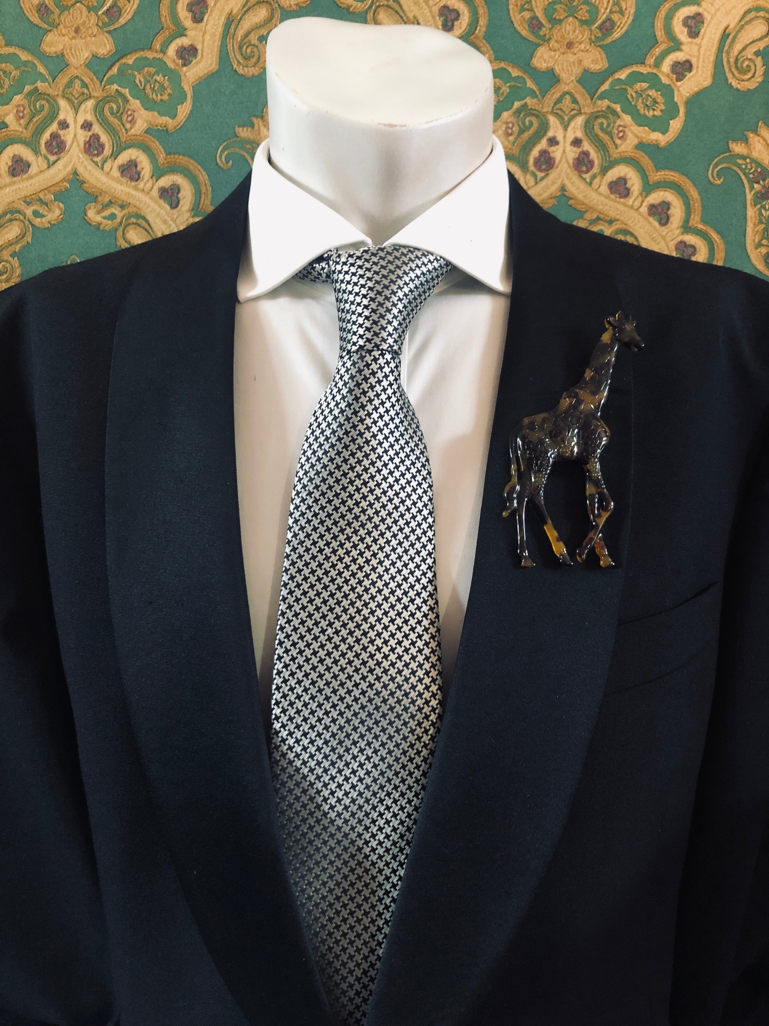 A Broochella Animalia Collection brown tortoiseshell giraffe brooch and men's lapel pin in Italian cellulose acetate worn on the lapel of a black suit jacket with a white shirt and houndstooth tie, styled on a mannequin against a green and gold damask wallpaper background.