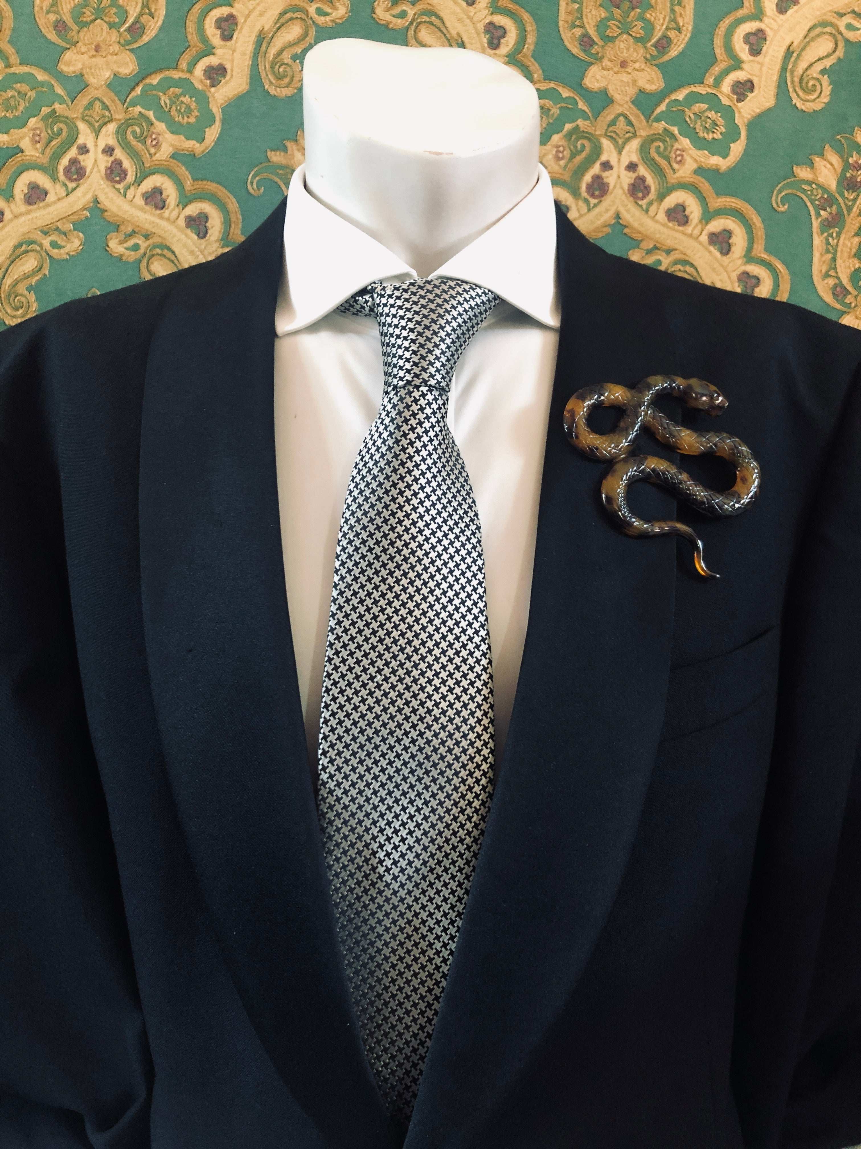A Broochella Animalia Collection beige tortoiseshell coiled snake brooch and men's lapel pin in Italian cellulose acetate worn on the lapel of a black suit jacket with a white shirt and houndstooth tie, styled on a mannequin against a green and gold damask wallpaper background.