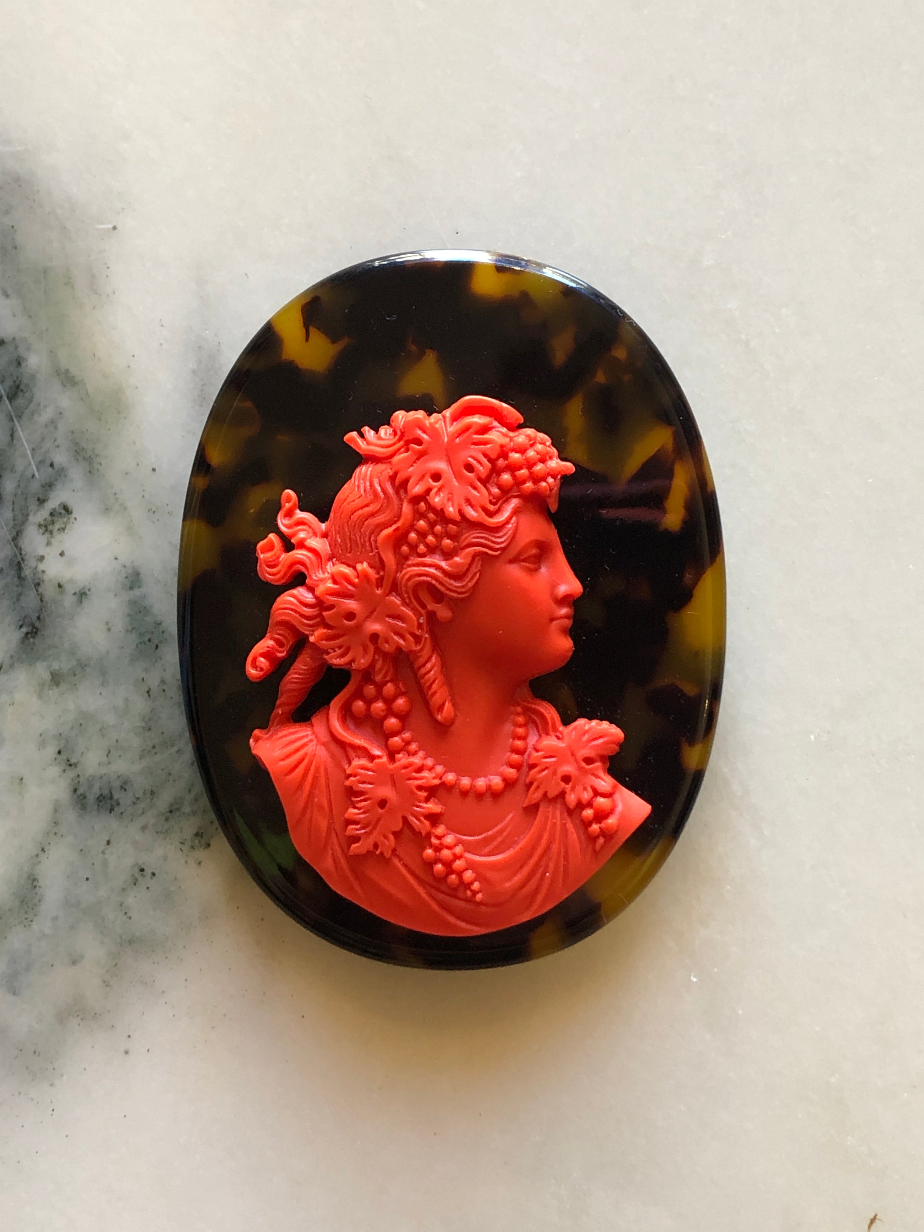 A Broochella La Bella Figura oval cameo brooch featuring a bright red classical profile bust of a woman with floral details, set against a dark brown and amber tortoiseshell-patterned background, displayed on a light marble surface.