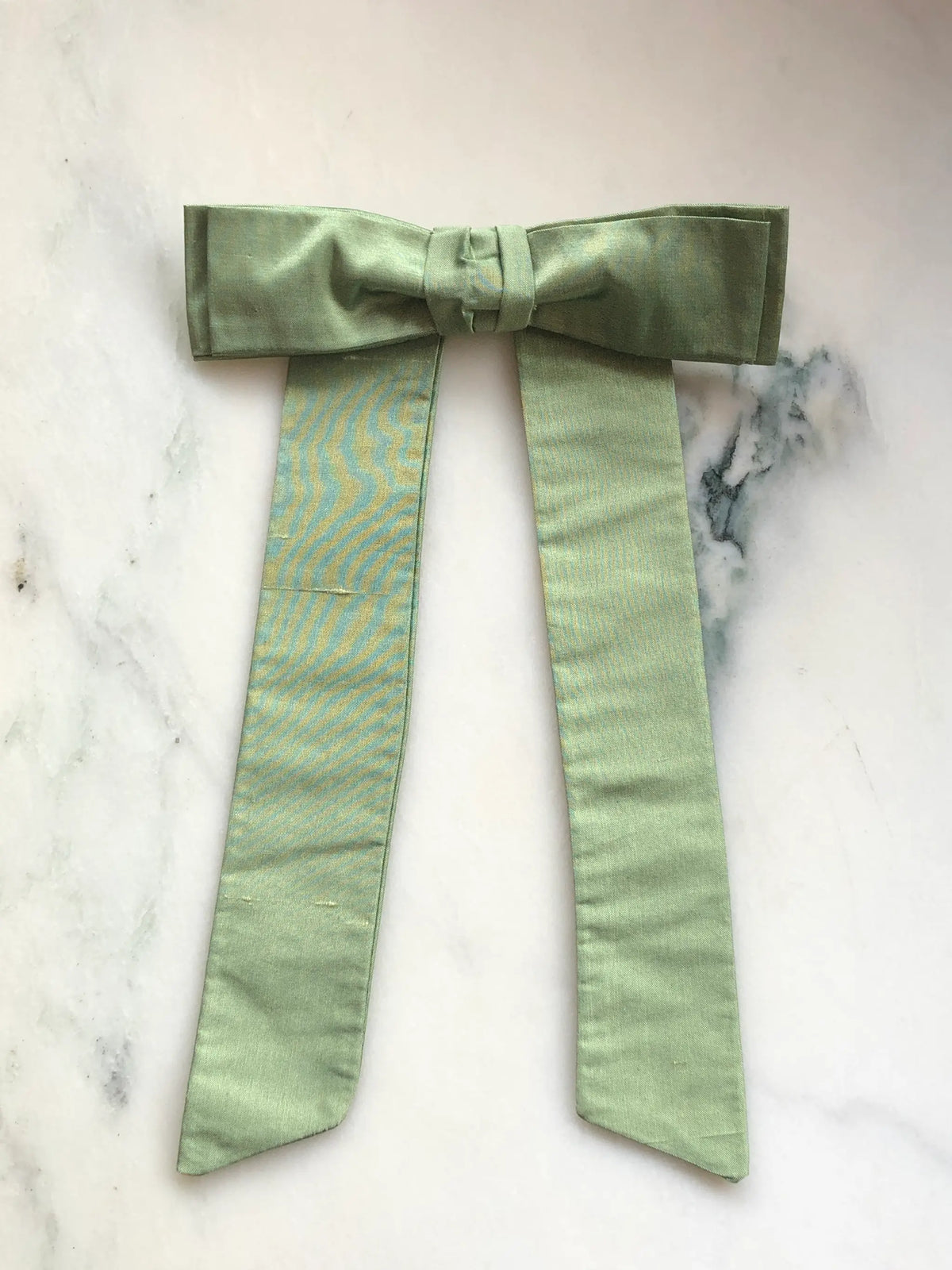 Handmade Broochella Alice Silk Bow Brooch in soft avocado green dupion silk, featuring an oversized designer bow with long elegant tails and a lustrous natural silk texture, photographed on white marble.