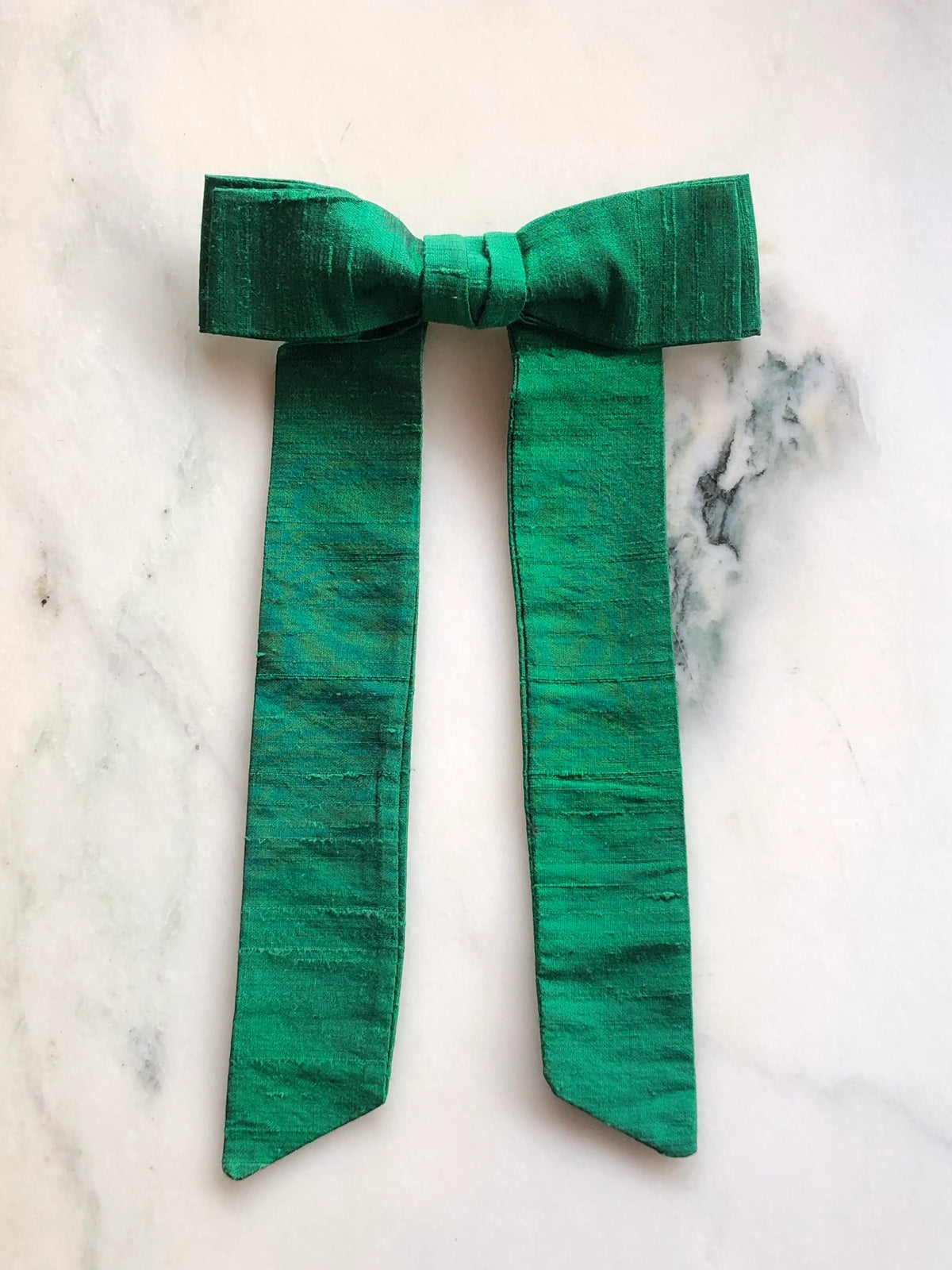 Handmade Broochella Alice Silk Bow Brooch in rich emerald green dupion silk, featuring an oversized designer bow with long elegant tails and lustrous natural silk texture, photographed on white marble.