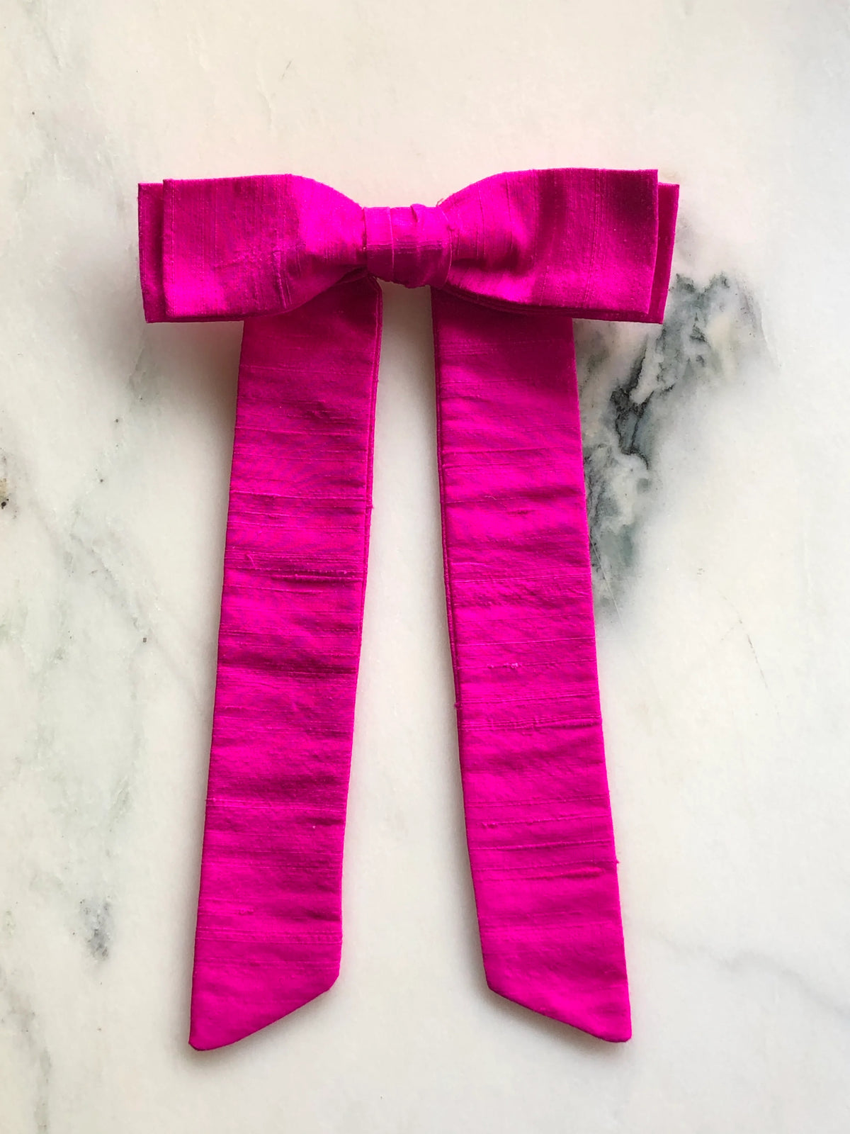 Handmade Broochella Alice Silk Bow Brooch in vibrant hot pink dupion silk, featuring an oversized designer bow with long elegant tails and lustrous natural silk texture, photographed on white marble.