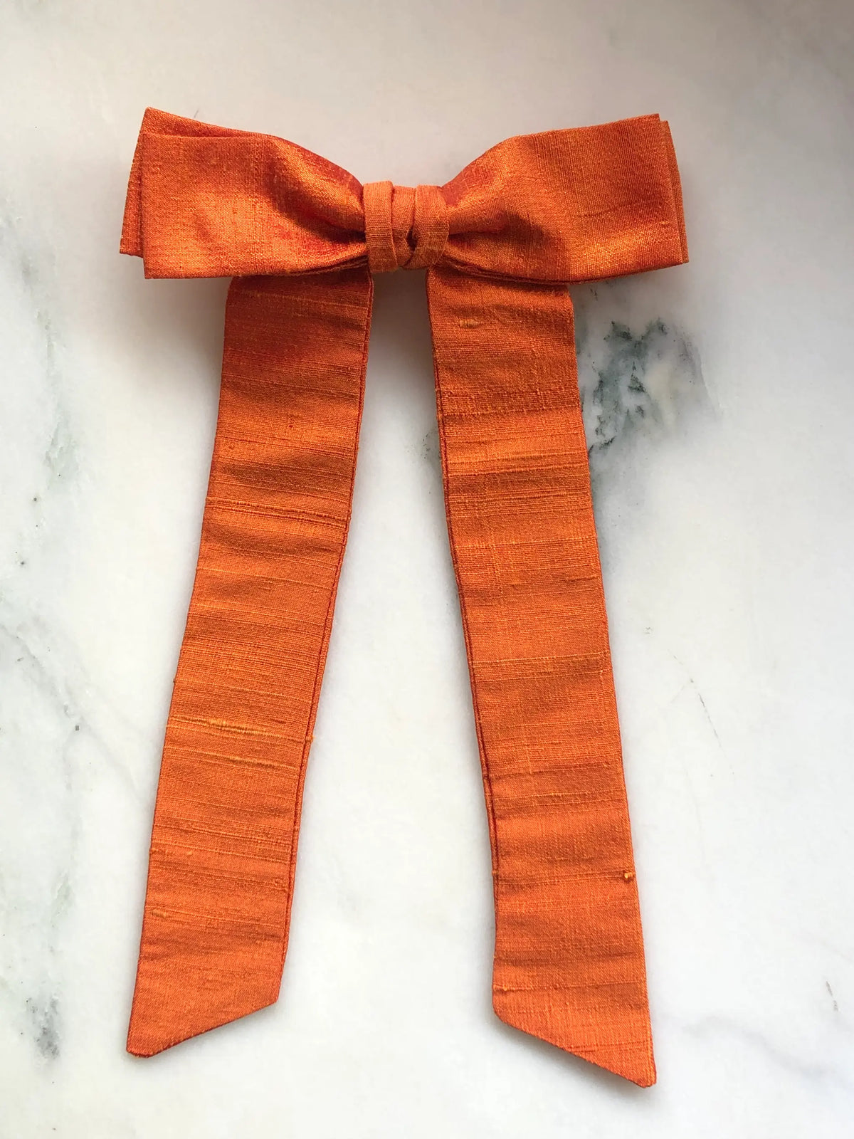 Handmade Broochella Alice Silk Bow Brooch in vibrant orange dupion silk, featuring an oversized designer bow with long elegant tails and natural silk texture, photographed on white marble.