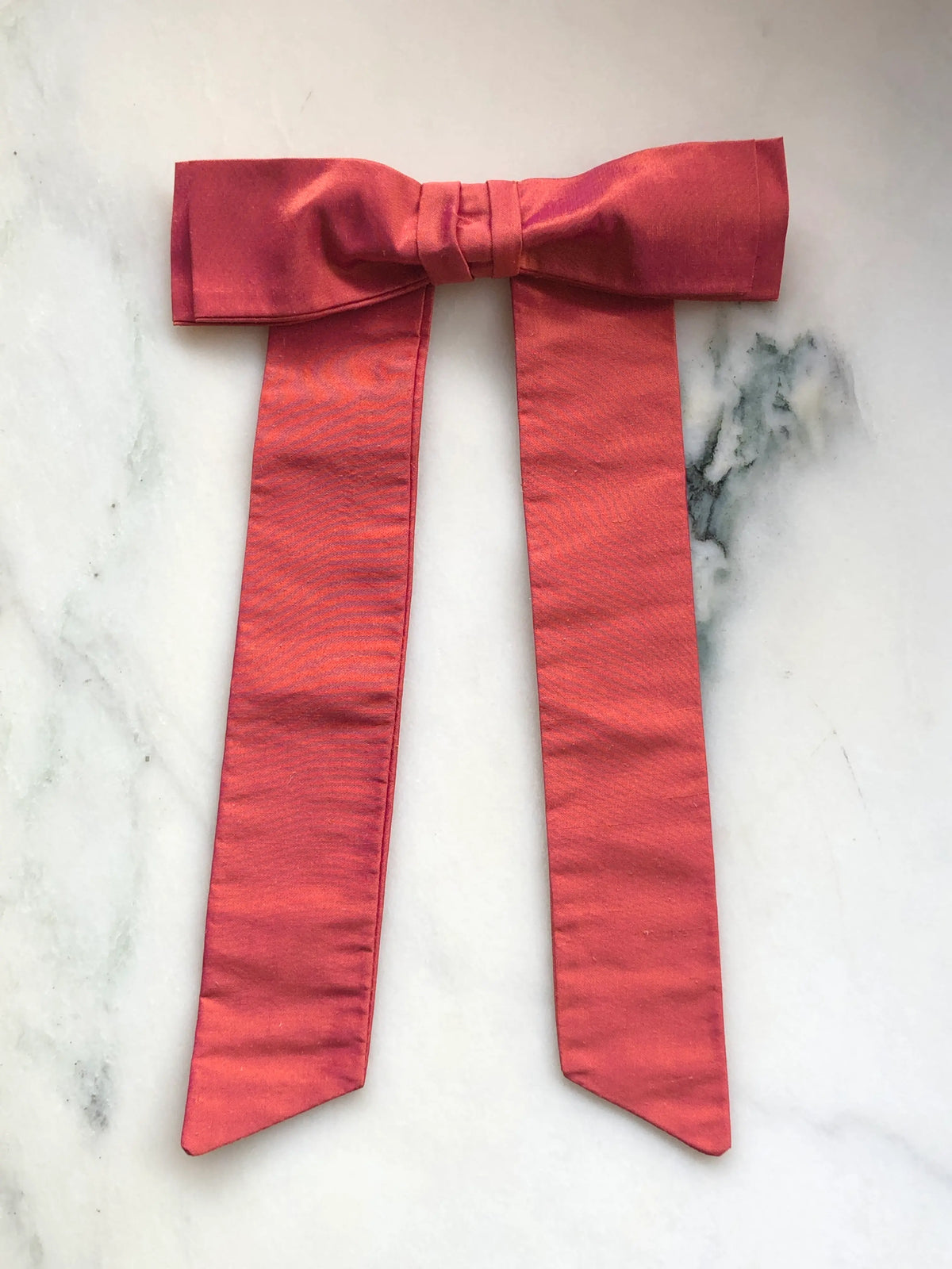 Handmade Broochella Alice Silk Bow Brooch in warm peach dupion silk, featuring an oversized designer bow with long elegant tails and a lustrous natural silk texture, photographed on white marble.