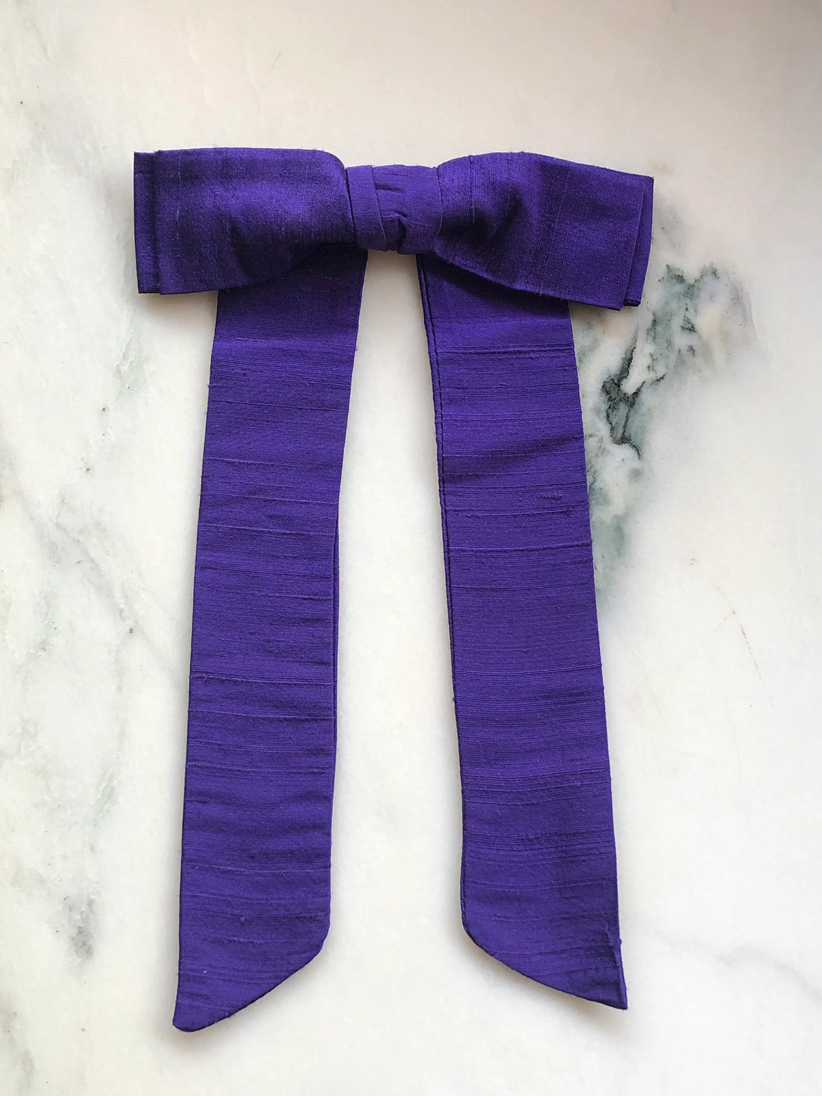 Handmade Broochella Alice Silk Bow Brooch in rich purple dupion silk, featuring an oversized designer bow with long elegant tails and natural silk texture, photographed on white marble.