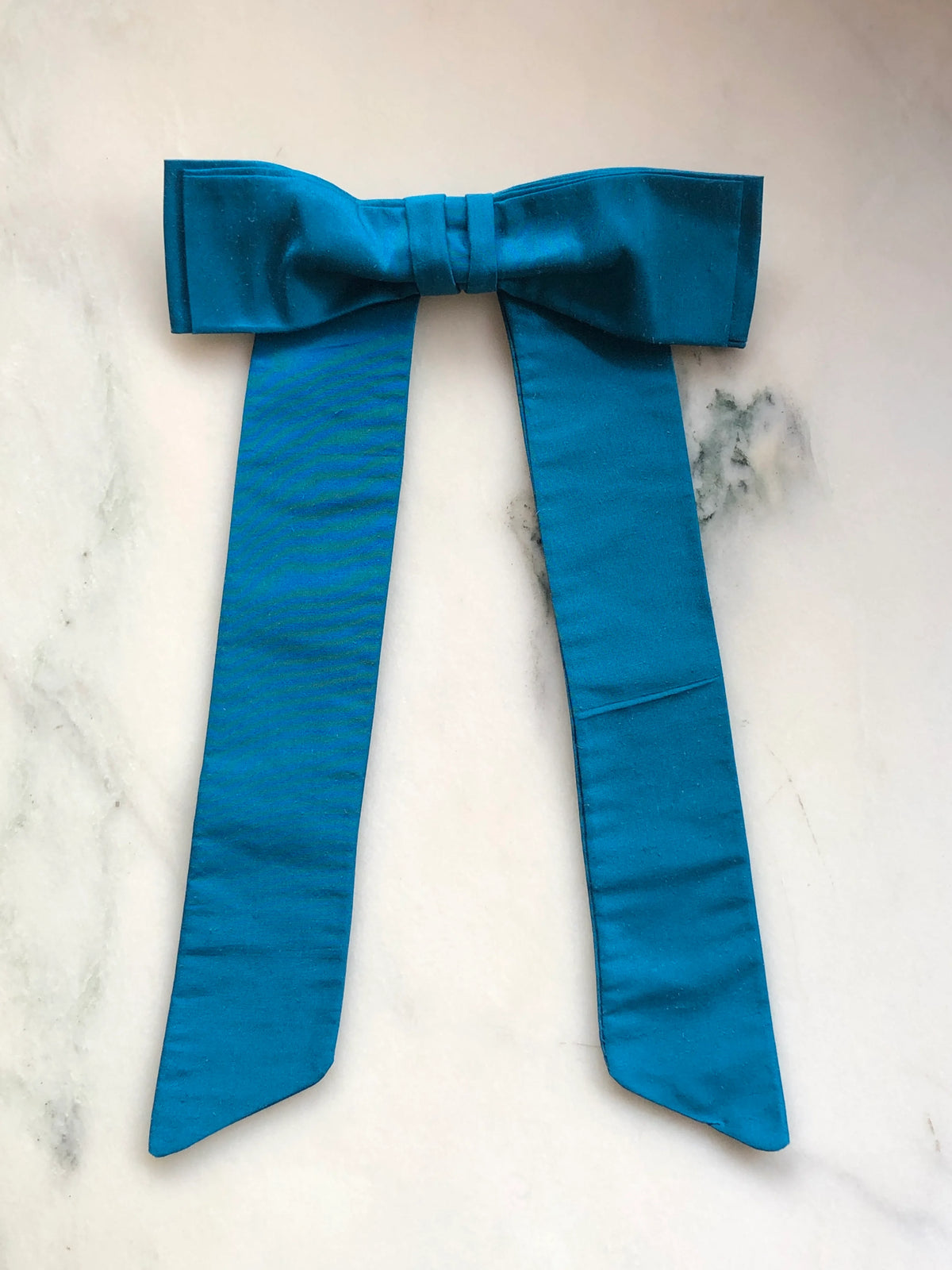 Handmade Broochella Men’s Alice Silk Bow Brooch in vibrant turquoise blue dupion silk, featuring an oversized designer bow with long tailored tails and a lustrous natural silk texture, photographed on white marble.