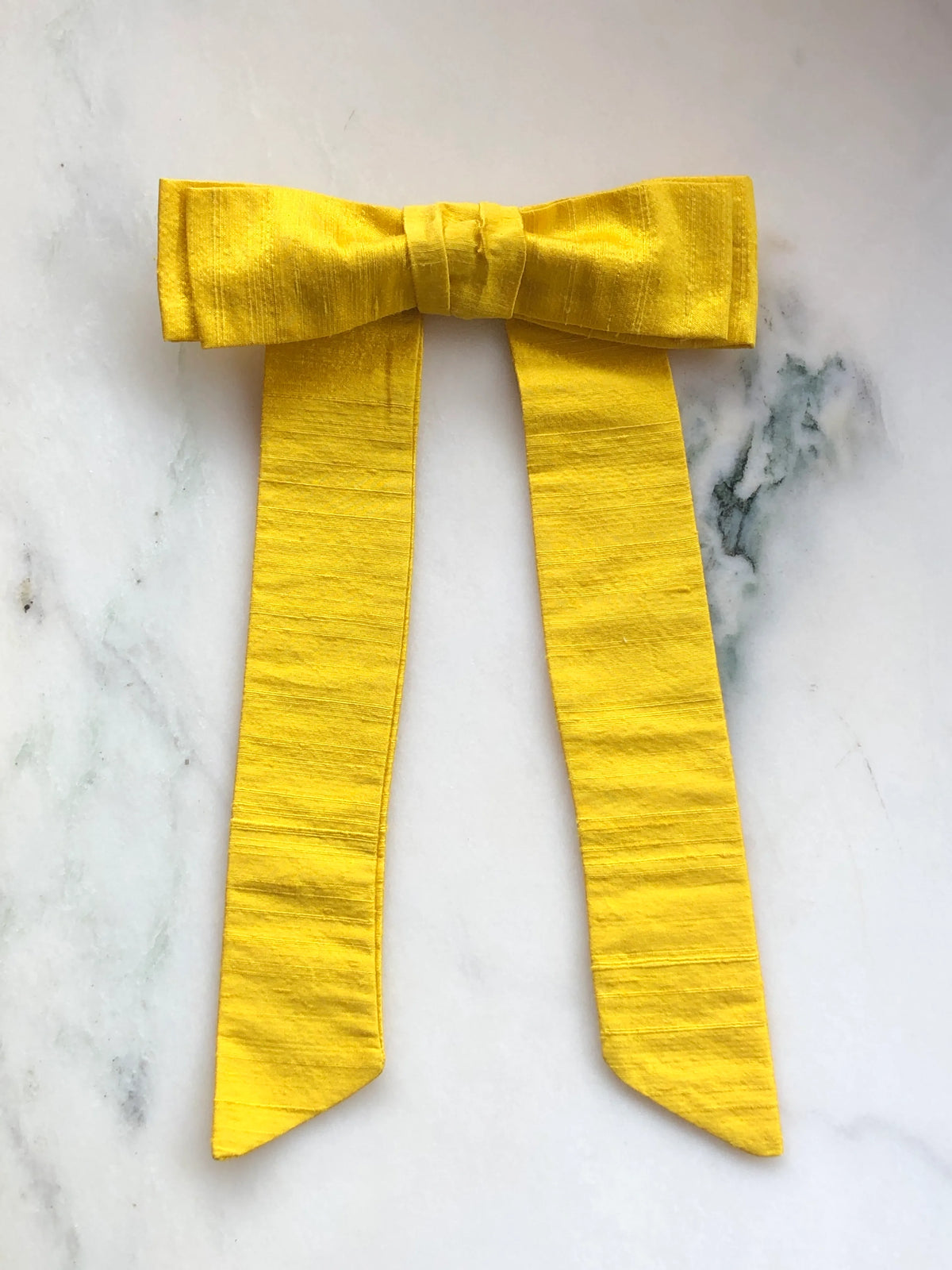 Handmade Broochella Alice Silk Bow Brooch in vibrant yellow dupion silk, featuring an oversized designer bow with long elegant tails and lustrous natural silk texture, photographed on white marble.