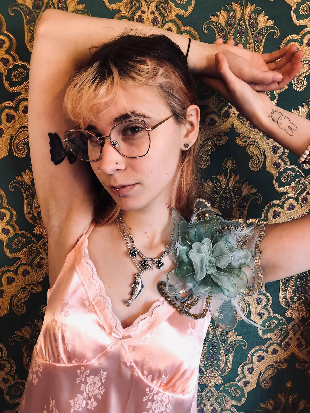 A person with glasses and tattoos poses against a dark green and gold damask patterned wallpaper, wearing a pale pink satin camisole and a Broochella Alstromeria brooch in green/blue.