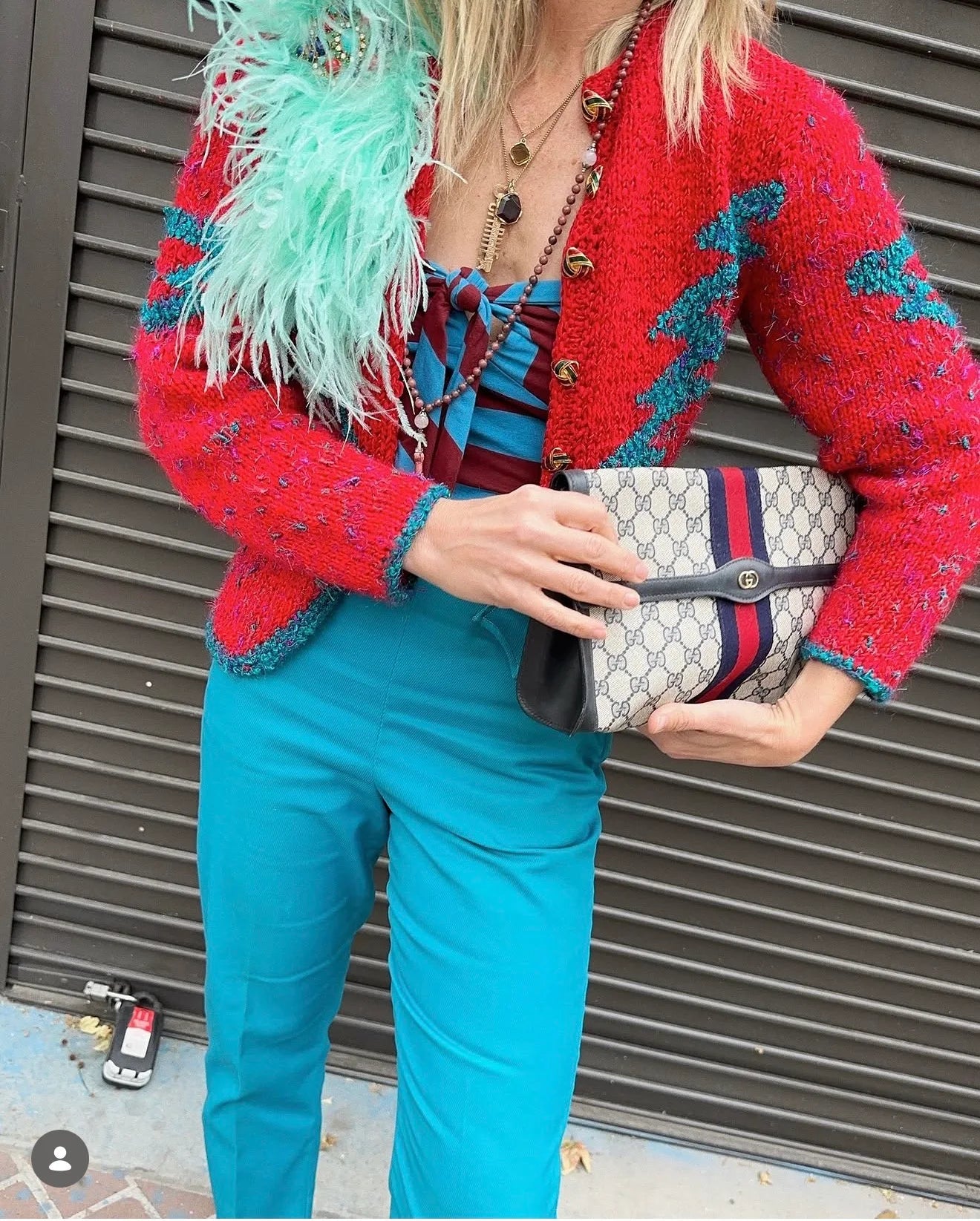 A person wearing a bright red Gucci tweed cardigan with turquoise feather accents, turquoise trousers, and holding a Gucci monogram clutch bag in front of a dark metal shutter. She is wearing the Broochella bejewelled handmade feather brooch in aqua.