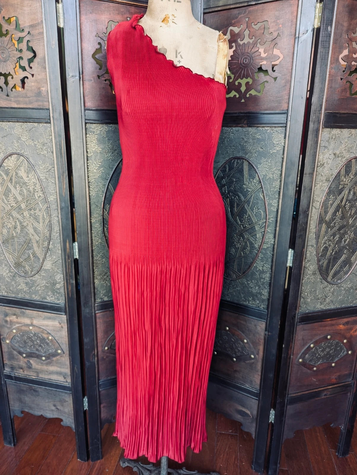 A Broochella l'idée bright red one-shoulder micro pleat dress with a scalloped neckline and a long pleated fringe hem, displayed on a mannequin torso in front of a dark wooden folding screen.