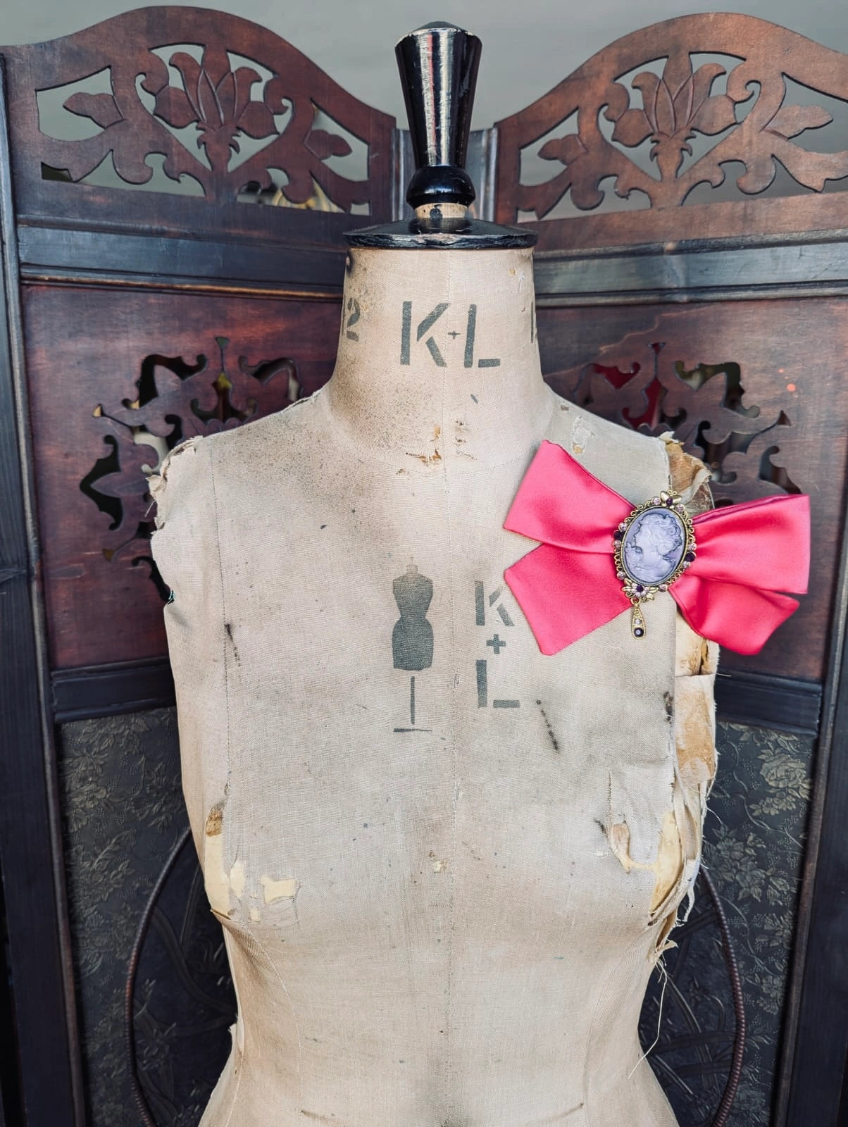 Broochella rosee camee pink handmade satin bow brooch with vintage cameo brooch embellishment. It is pinned to a vintage mannequin. The background is a decorative brown wooden screen.
