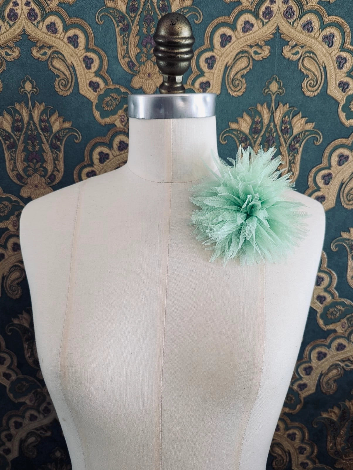 Broochella monello handmade tulle urchin brooch in mint. The brooch is pinned on a mannequin. The background is green and gold damask wallpaper.