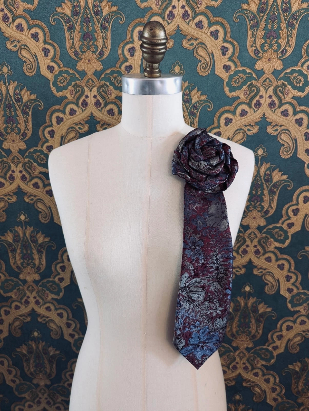 Broochella Fuente stefano vintage up-cycled vintage handcrafted rosette tie in burgundy and blue damask. Pinned on a mannequin. The background is green and gold damask wallpaper