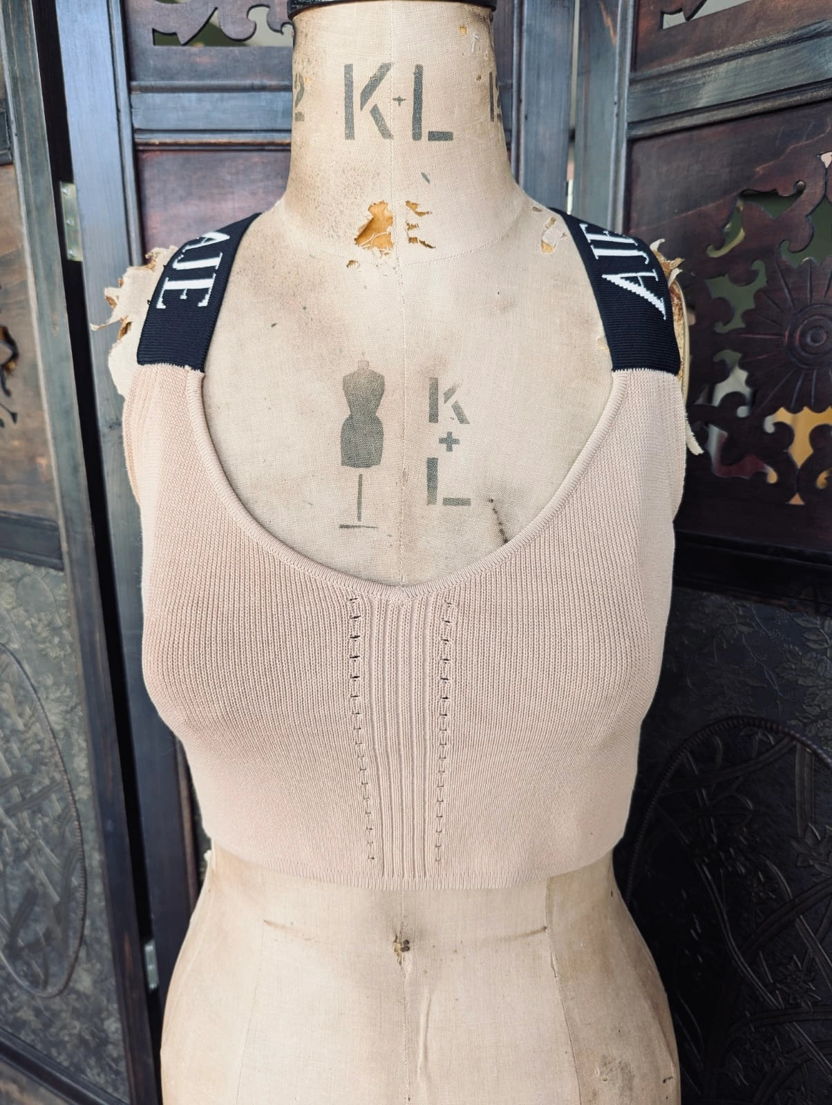 A Broochella Aje beige ribbed knit scoop-neck crop top displayed on a vintage wooden mannequin torso. The top features black shoulder straps with white lettering, set against a dark wooden background.