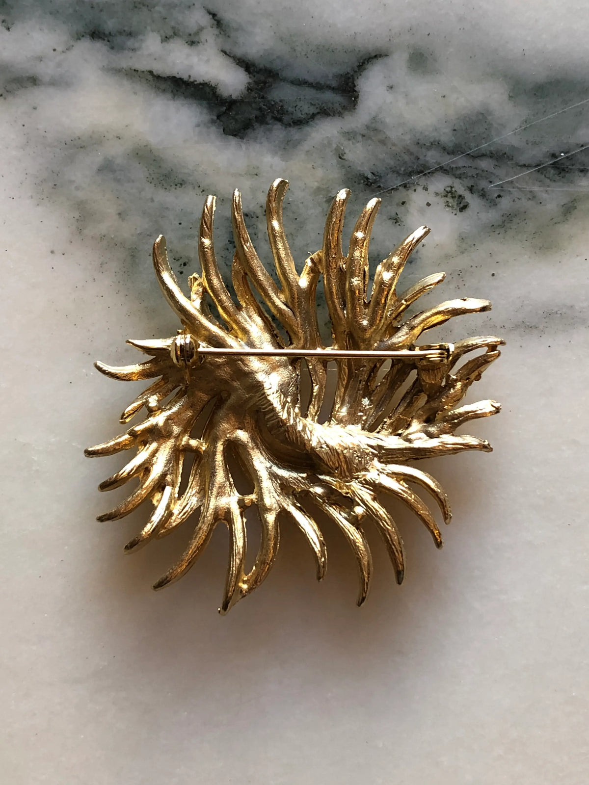The back view of Broochella Alessandra Vintage Gold Swirl Brooch featuring sculptural textured gold-tone fronds arranged in a circular swirling design on a cream and grey marble background.
