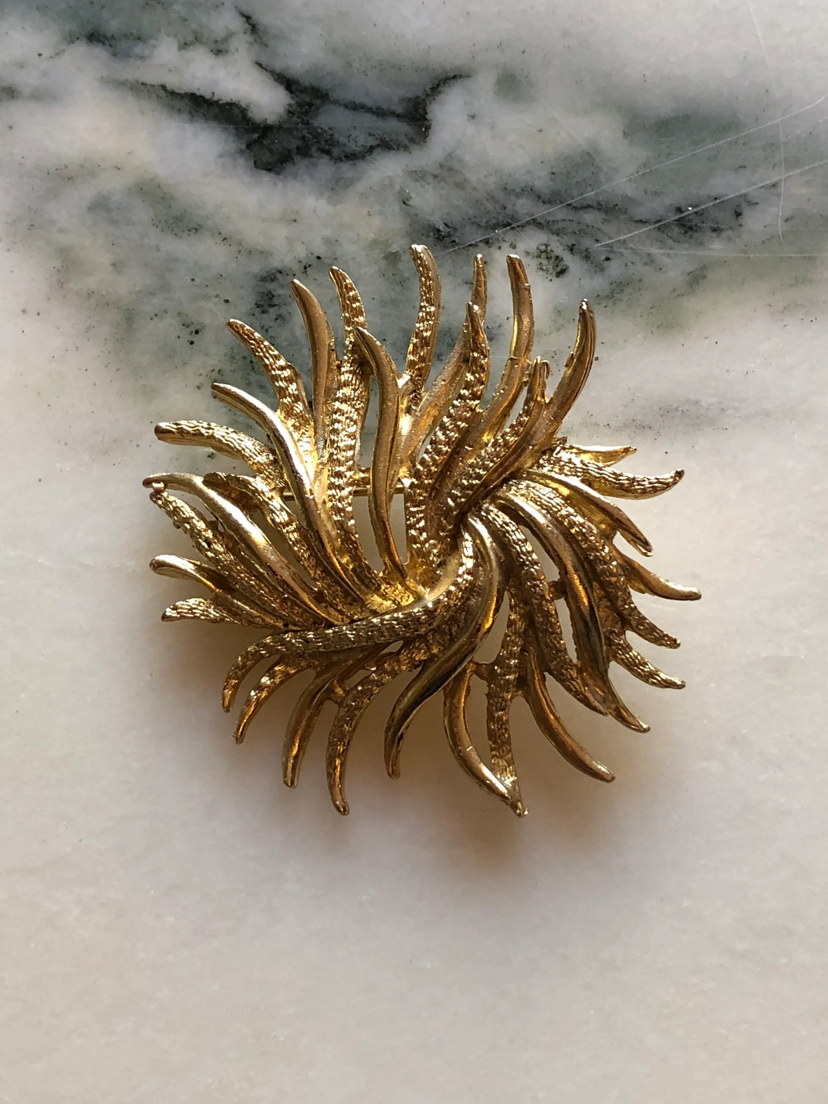 Broochella Alessandra Vintage Gold Swirl Brooch featuring sculptural textured gold-tone fronds arranged in a circular swirling design on a cream and grey marble background.
