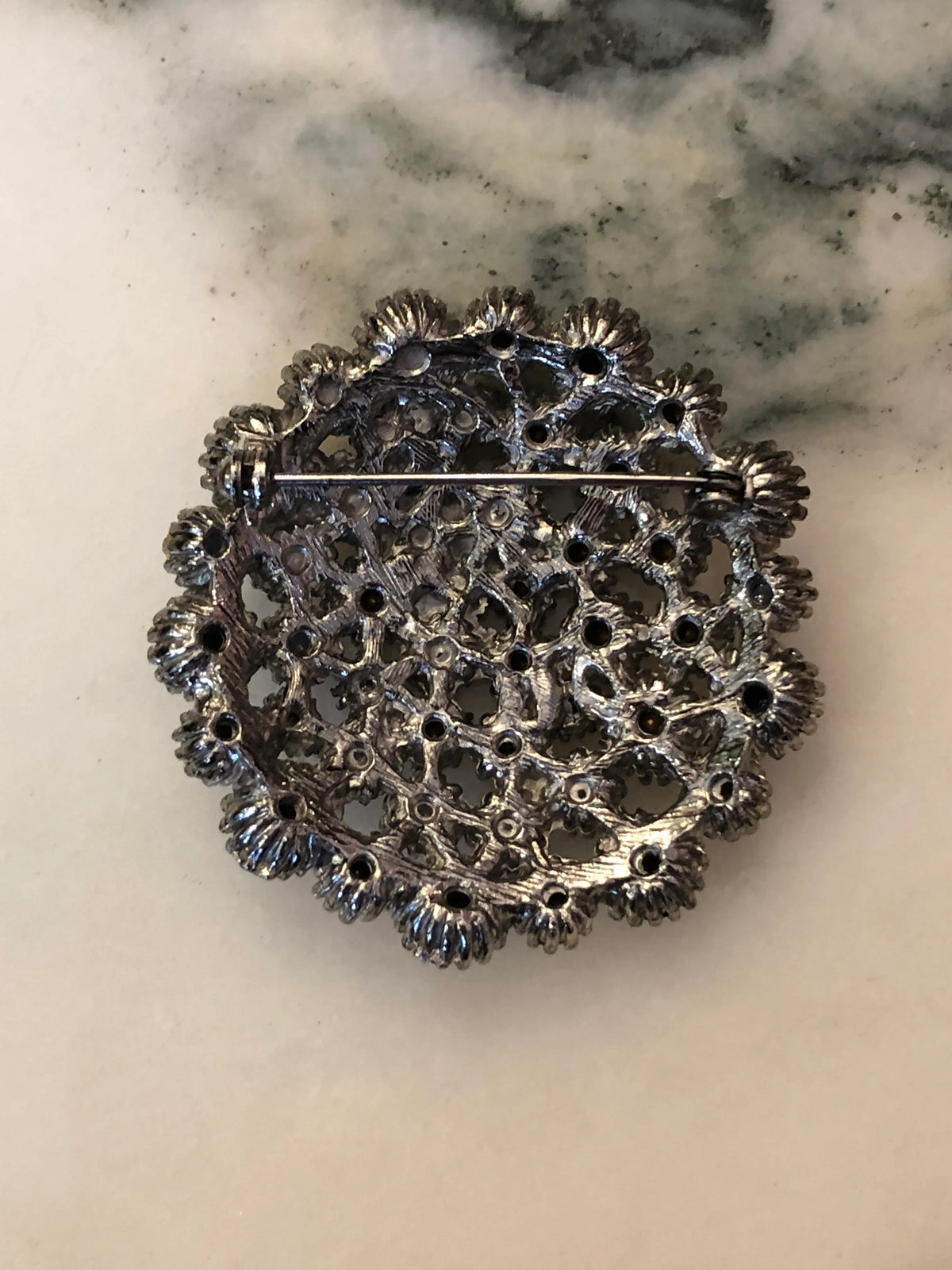 The back view Broochella Alessia Vintage Black & Crystal Brooch featuring clear crystals and jet-black stones in an ornate silver-tone circular setting on a cream and grey marble background.