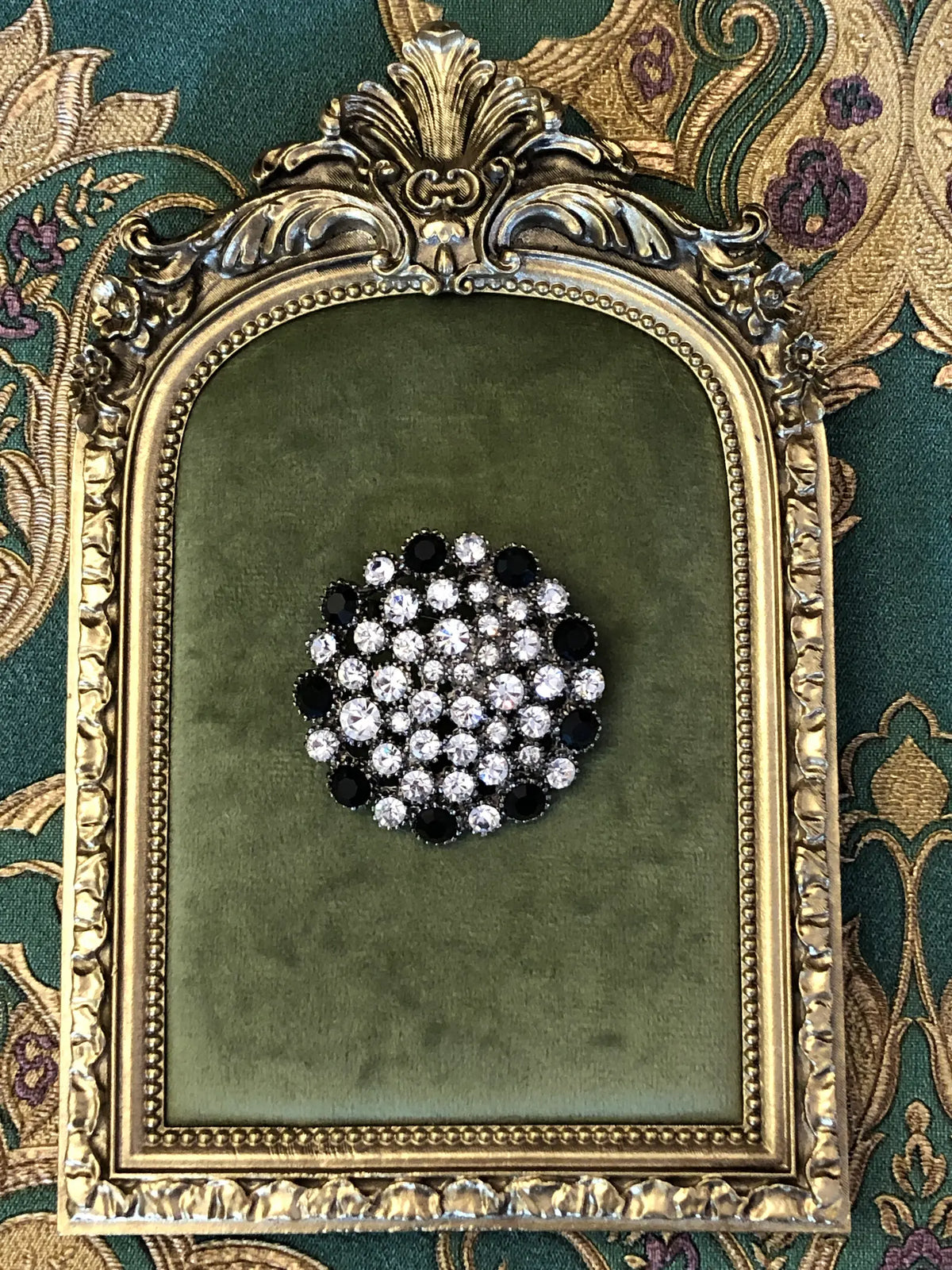 Broochella Alessia Vintage Black & Crystal Brooch featuring clear crystals and jet-black stones in an ornate silver-tone circular setting. It is pictured on a green velvet tray with an ornate gold frame. The background is green and gold damask wallpaper
