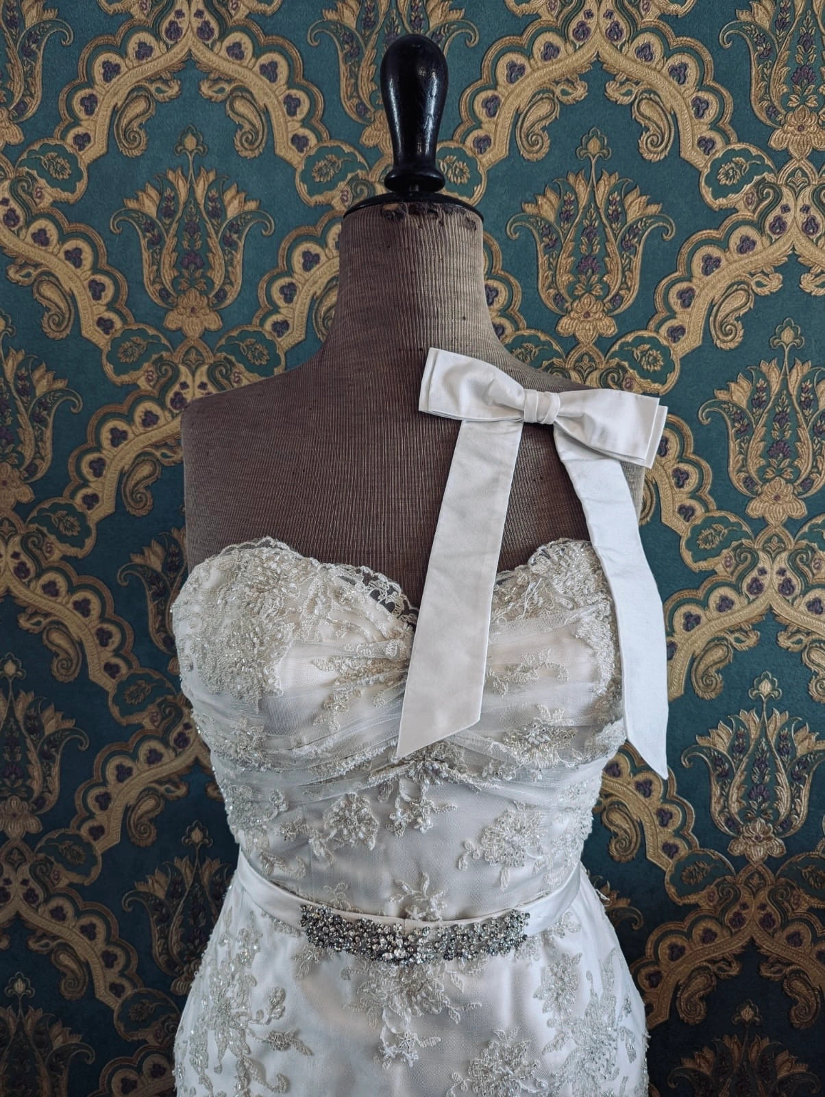Broochella bridal handmade silk bow in ivory. Pinned on a mannequin that is wearing a white, beaded strapless wedding dress. The background is green and gold damask wallpaper. 