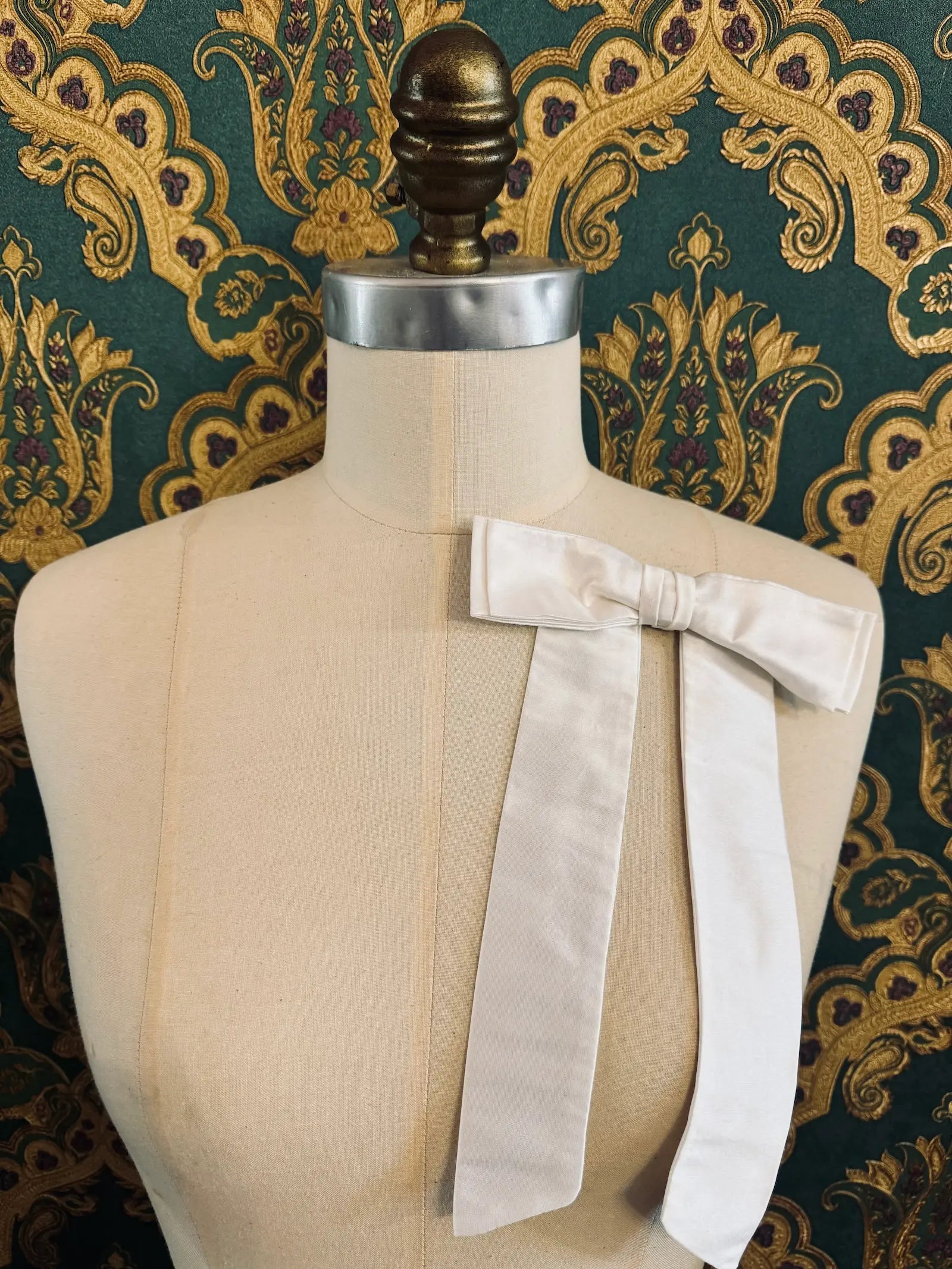 Broochella Alice silk handmade bow brooch in ivory. Pictured on a mannequin.  The background is green and gold damask wallpaper.