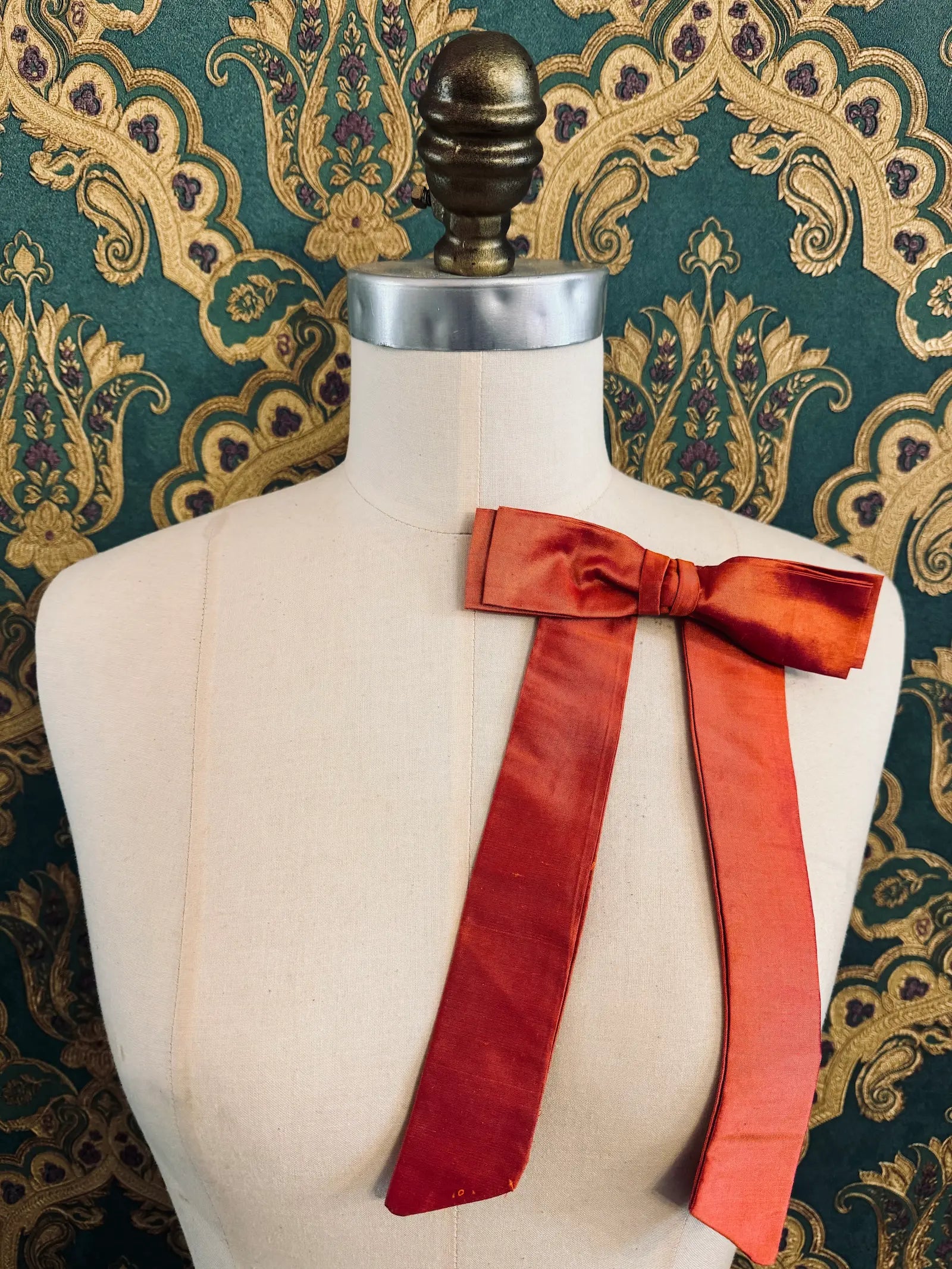 Broochella Alice silk handmade bow brooch in peach. Pictured on a mannequin.  The background is green and gold damask wallpaper.