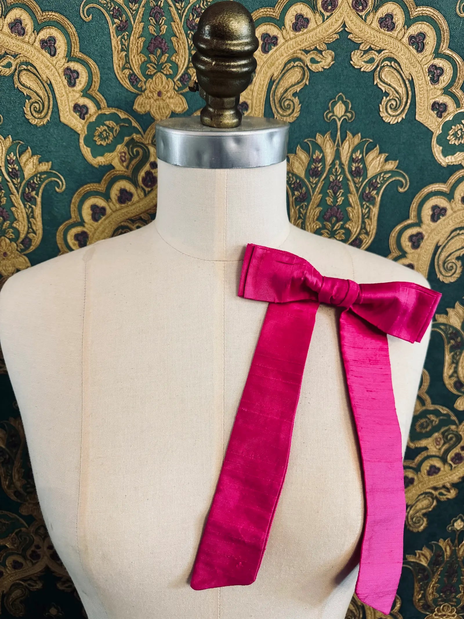 Broochella Alice silk handmade bow brooch in pink. Pictured on a mannequin.  The background is green and gold damask wallpaper.