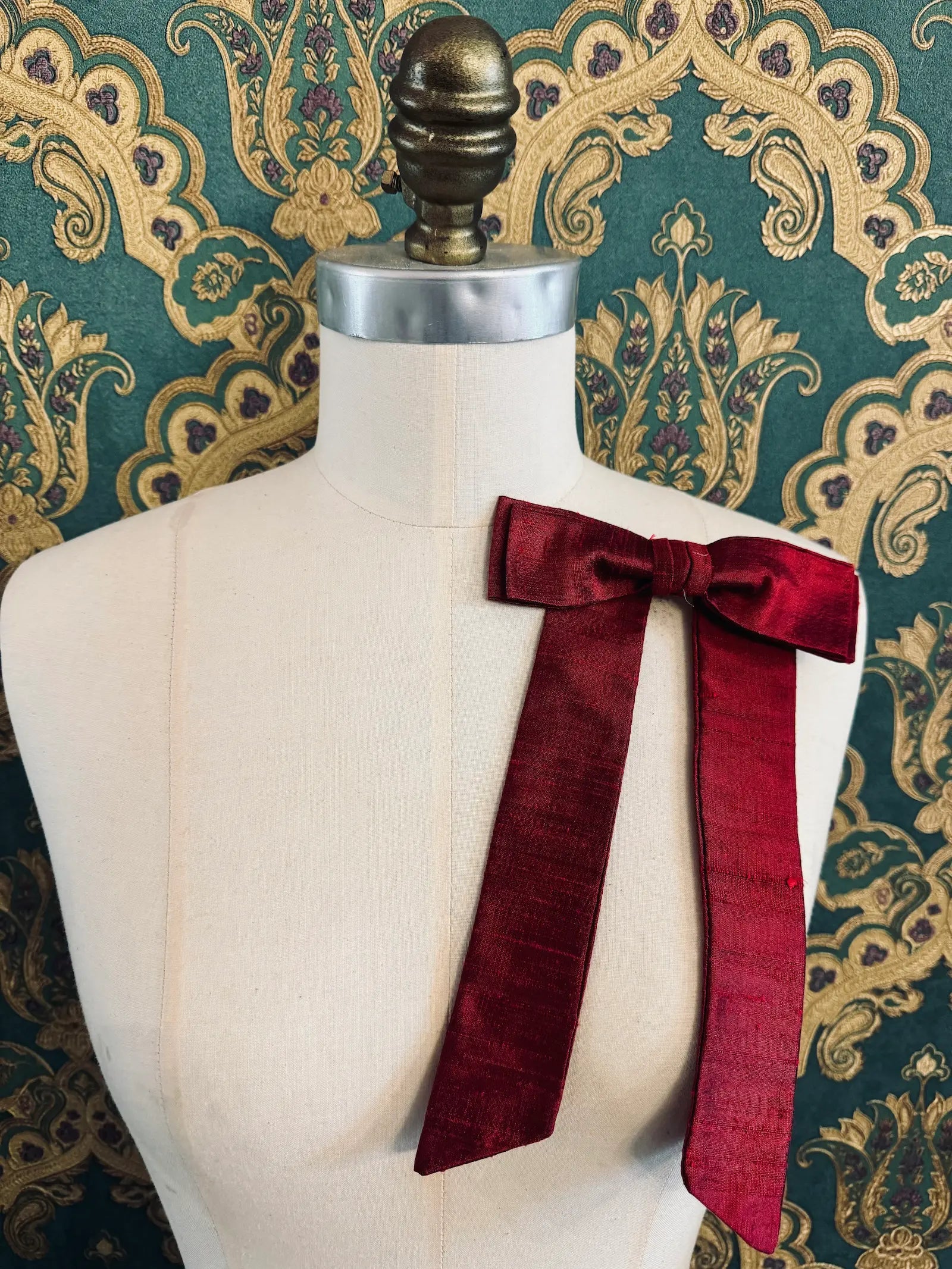 Broochella Alice silk handmade bow brooch in red. Pictured on a mannequin.  The background is green and gold damask wallpaper.