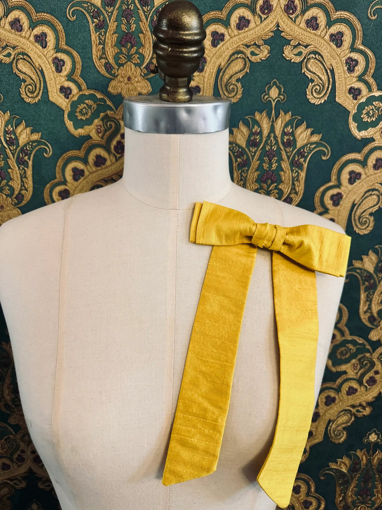Broochella Alice silk handmade bow brooch in yellow. Pictured on a mannequin.  The background is green and gold damask wallpaper.