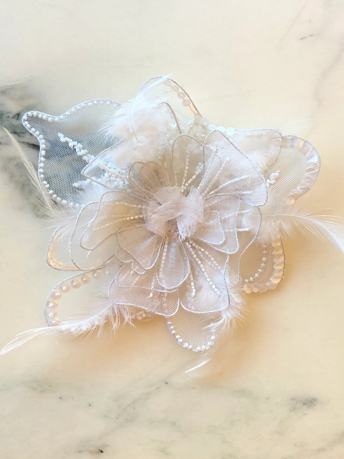 Broochella Alstroemeria Bridal Brooch Small featuring layered white mesh petals with intricate white beadwork, sequins and delicate feather accents, photographed on a pale cream marble background.