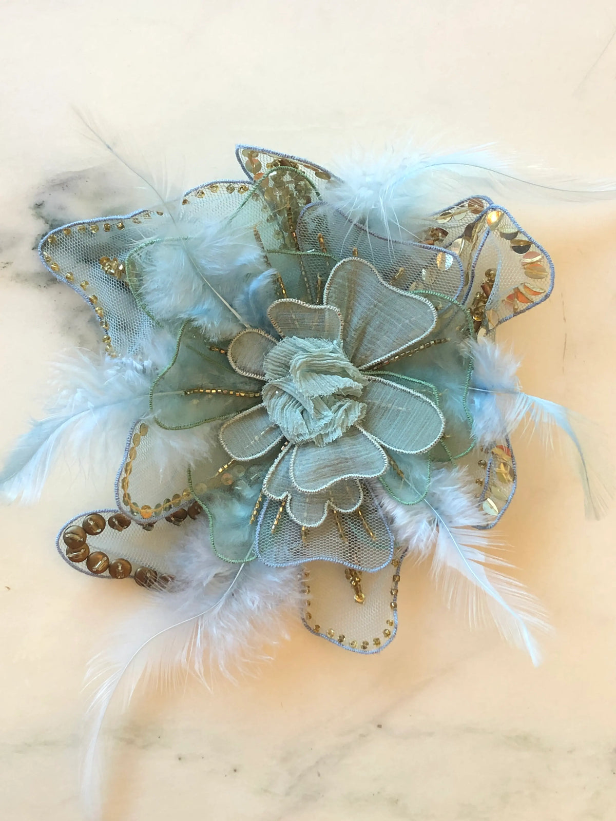 Broochella Alstroemeria Hand-Beaded Flower Brooch - Small in soft blue, featuring layered sheer organza petals embellished with gold sequins, beadwork and delicate pale blue feather accents on a cream marble background.