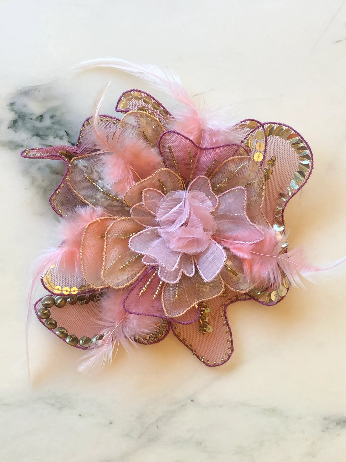 Broochella Men's Alstroemeria Hand-Beaded Flower Brooch - Small in soft pink, featuring layered sheer organza petals embellished with gold sequins and beads, purple edging and delicate pink feather accents on a cream marble background.